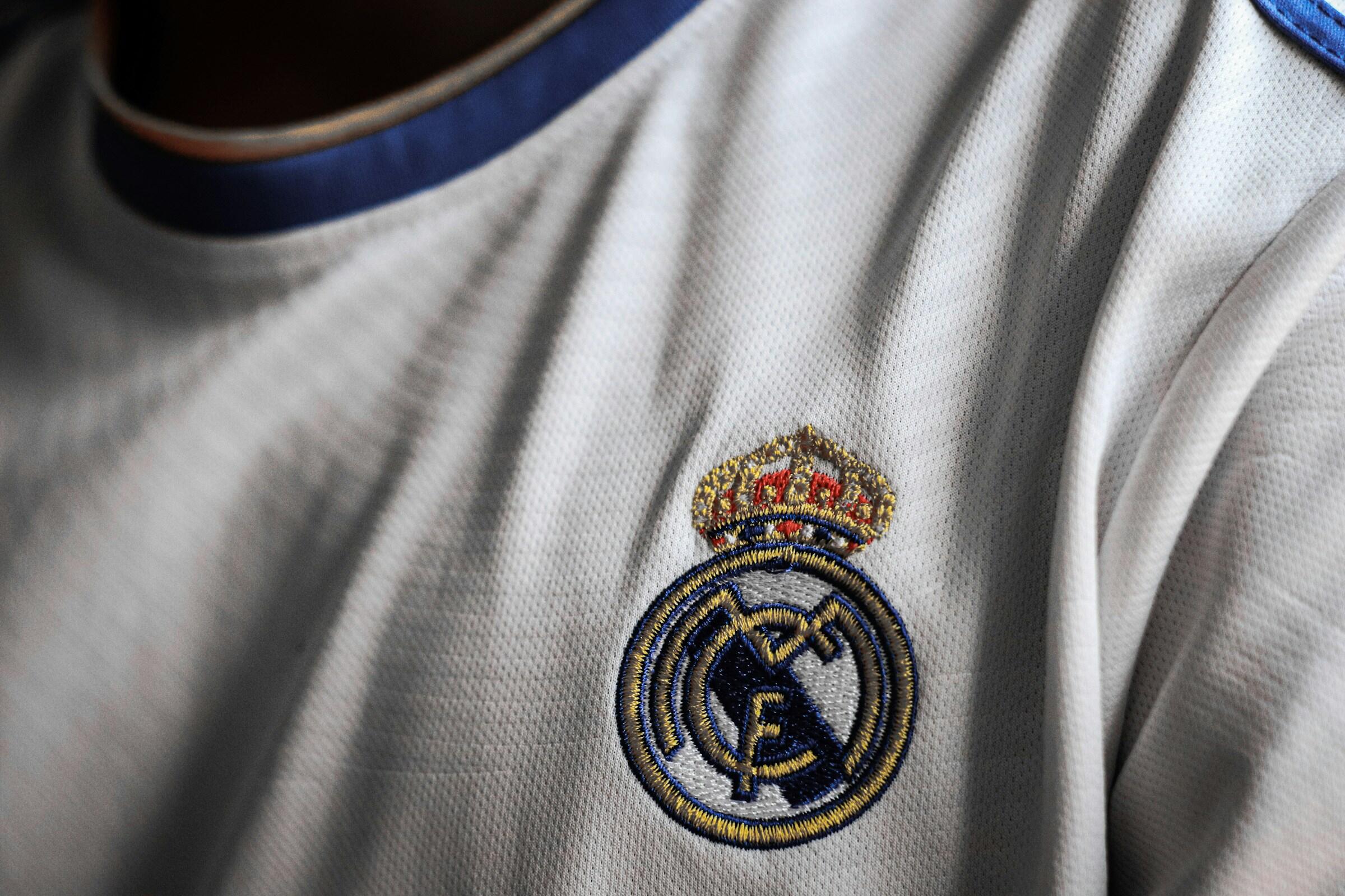 Primer plano del hombro de una persona que lleva una camiseta blanca del Real Madrid, con el logotipo del club bordado con una corona.