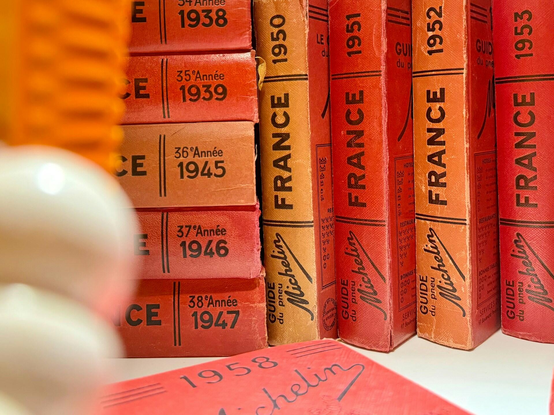 Primer plano de unas guías Michelin antiguas, de color rojo y naranja, que datan de entre 1938 y 1958, colocadas en una estantería. Los lomos desgastados transmiten una sensación de nostalgia e historia.