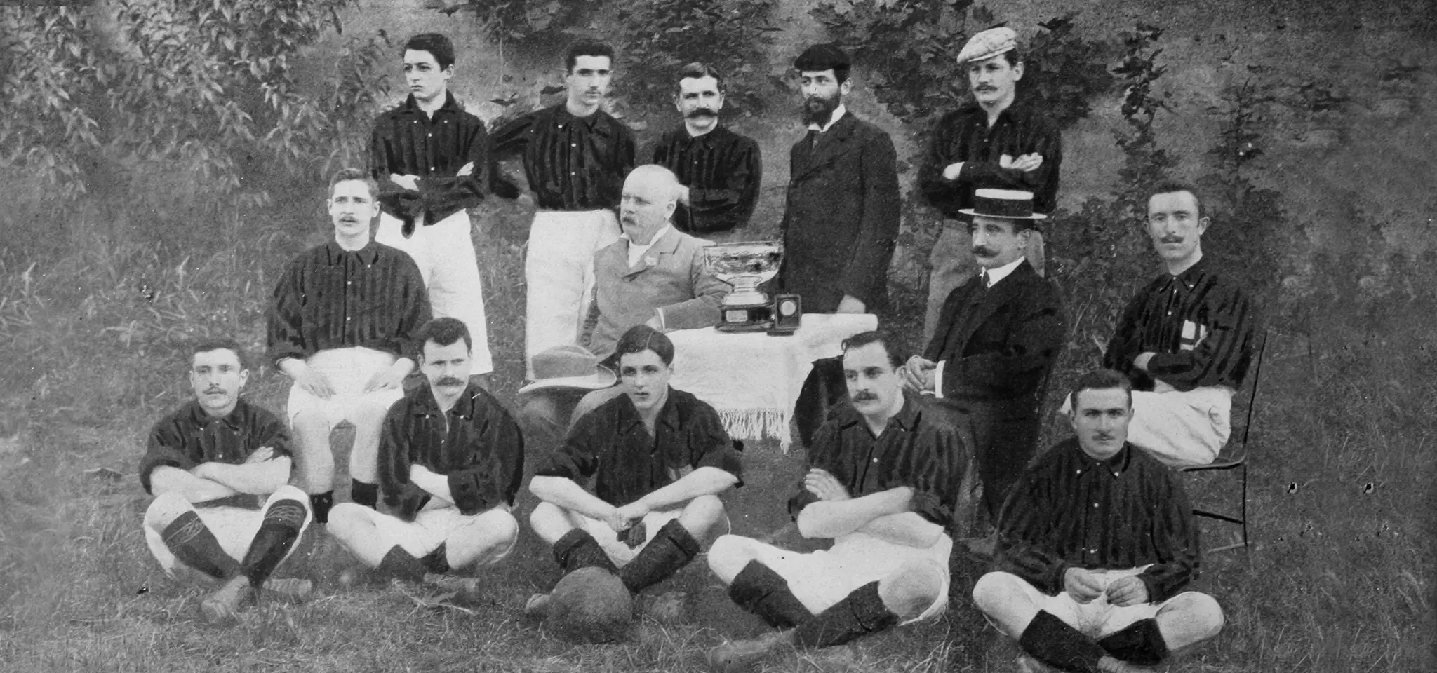 Foto histórica en blanco y negro del equipo de futbol ac milan con jugadores vestidos con camisetas a rayas, posando con un trofeo sobre una mesa al aire libre 