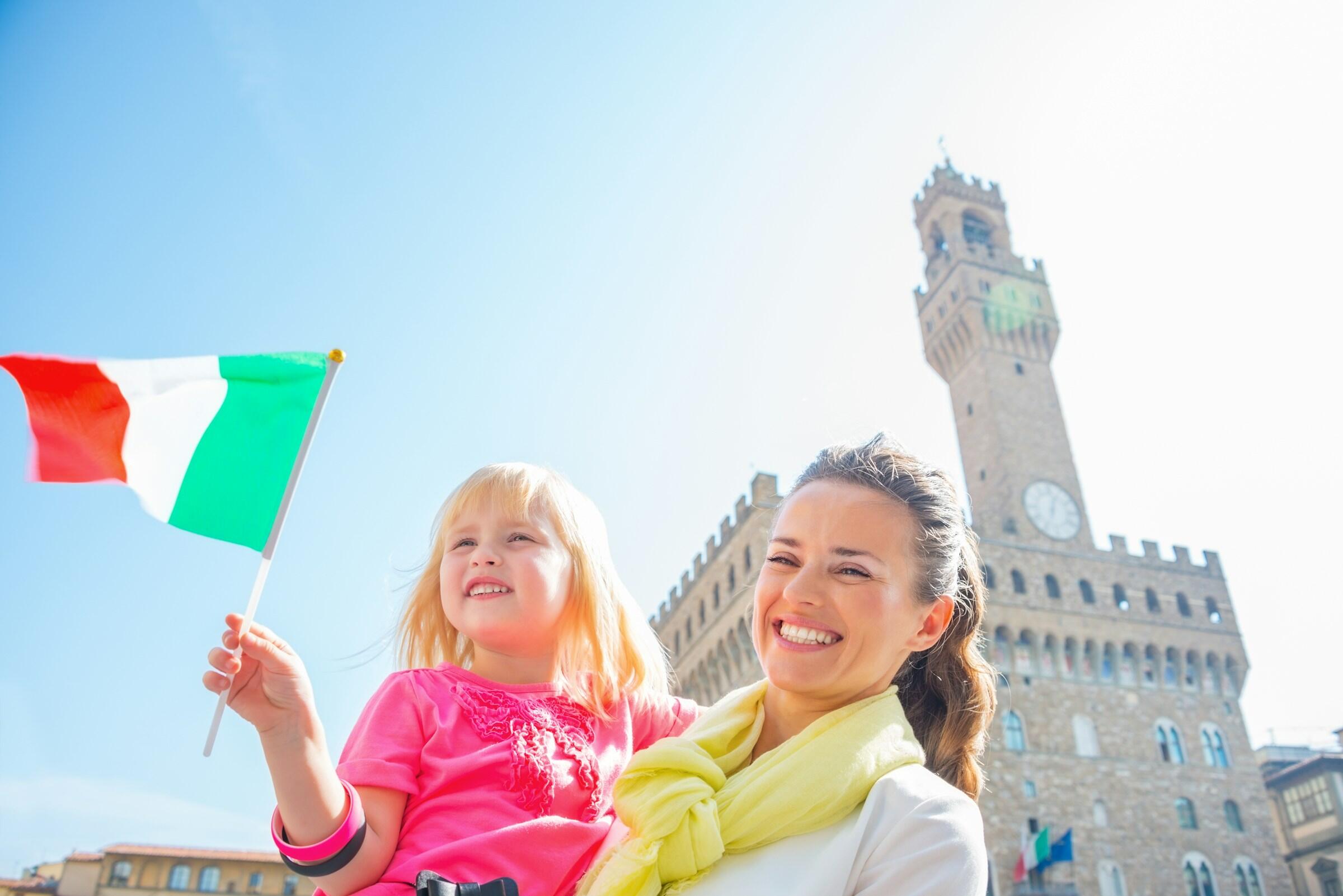 Una mujer sostiene a una niña que ondea una bandera italiana contra un cielo brillante, con el Palazzo Vecchio alzándose al fondo.
