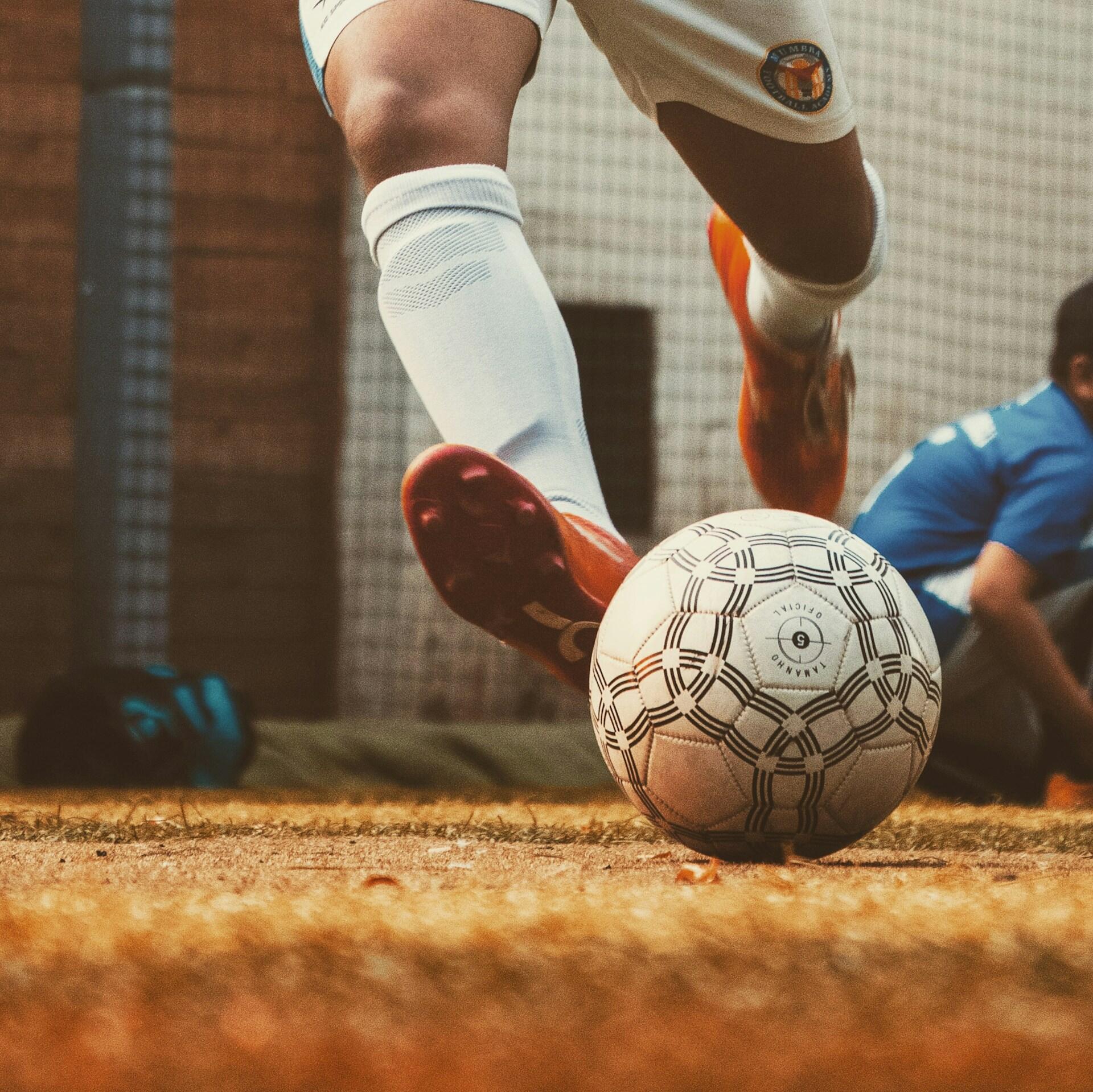 Primer plano de la pierna de un futbolista en movimiento, con calcetines blancos y botas naranjas, a punto de chutar un balón estampado en un campo de césped. Un momento lleno de acción.