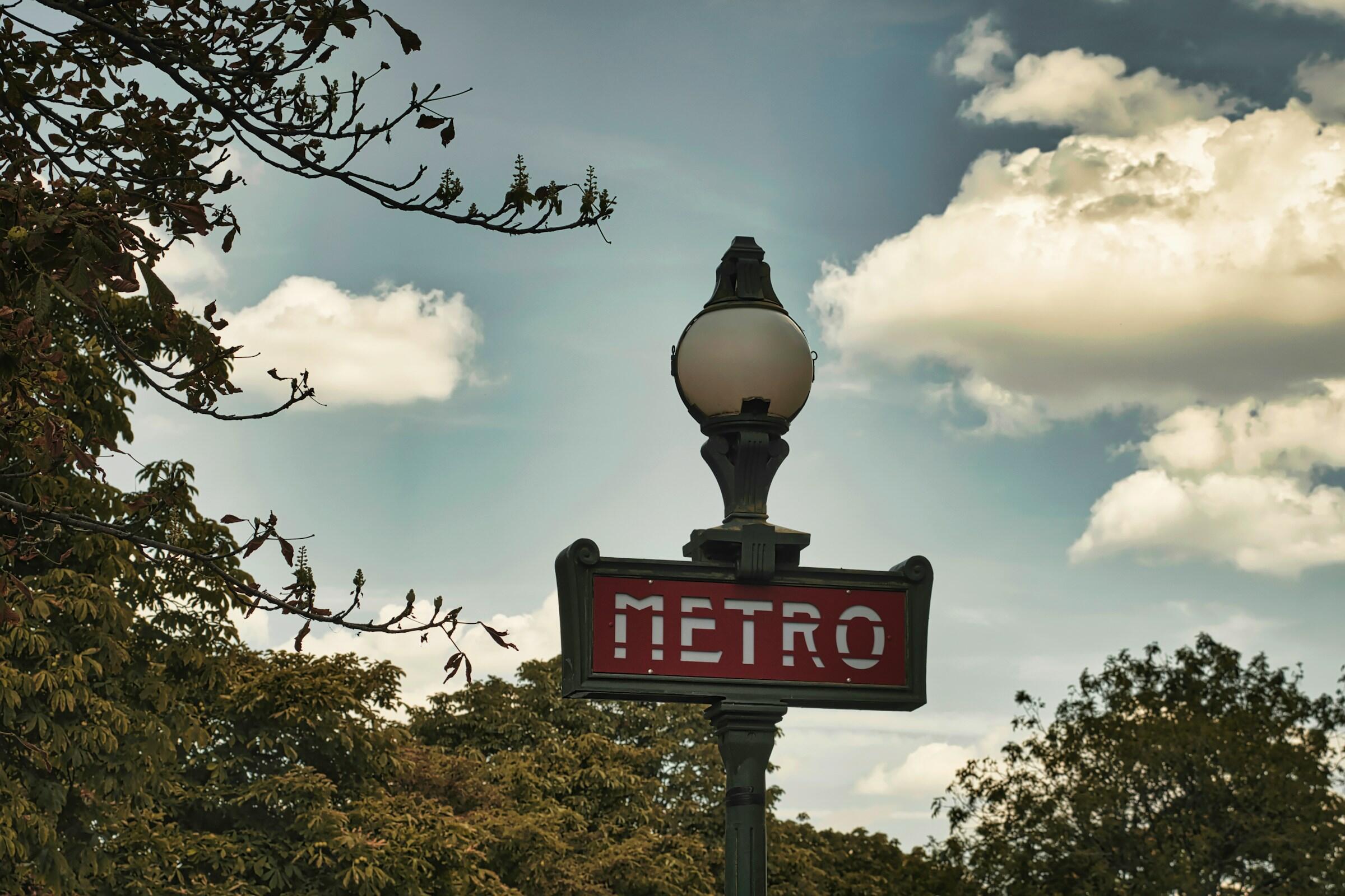 Una señal de metro vintage con una farola decorativa, rodeada de ramas de árbol y un cielo nublado.