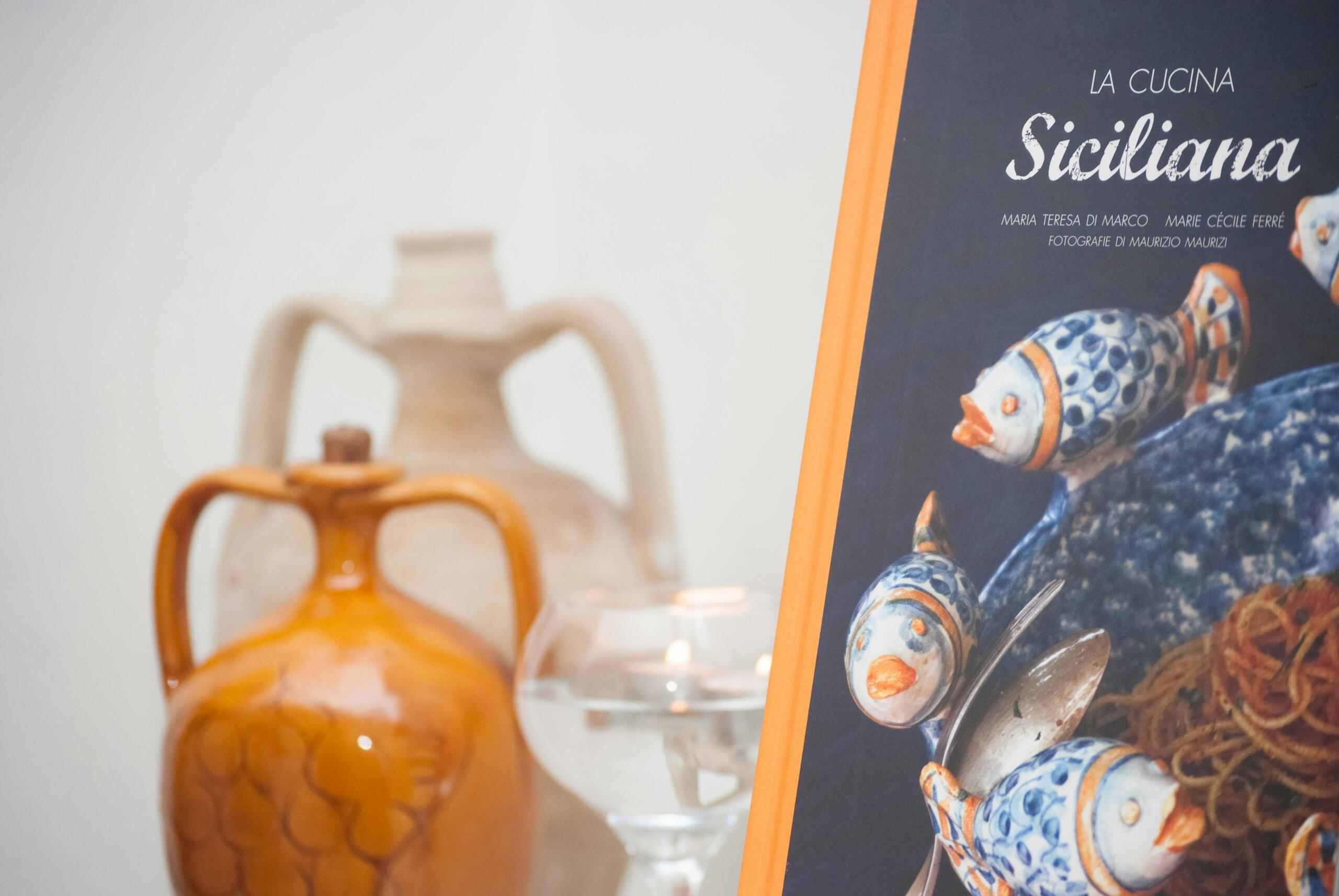 Un libro de cocina naranja titulado «La Cucina Siciliana» con coloridas ilustraciones de peces, junto a cerámica decorativa y un vaso de agua.