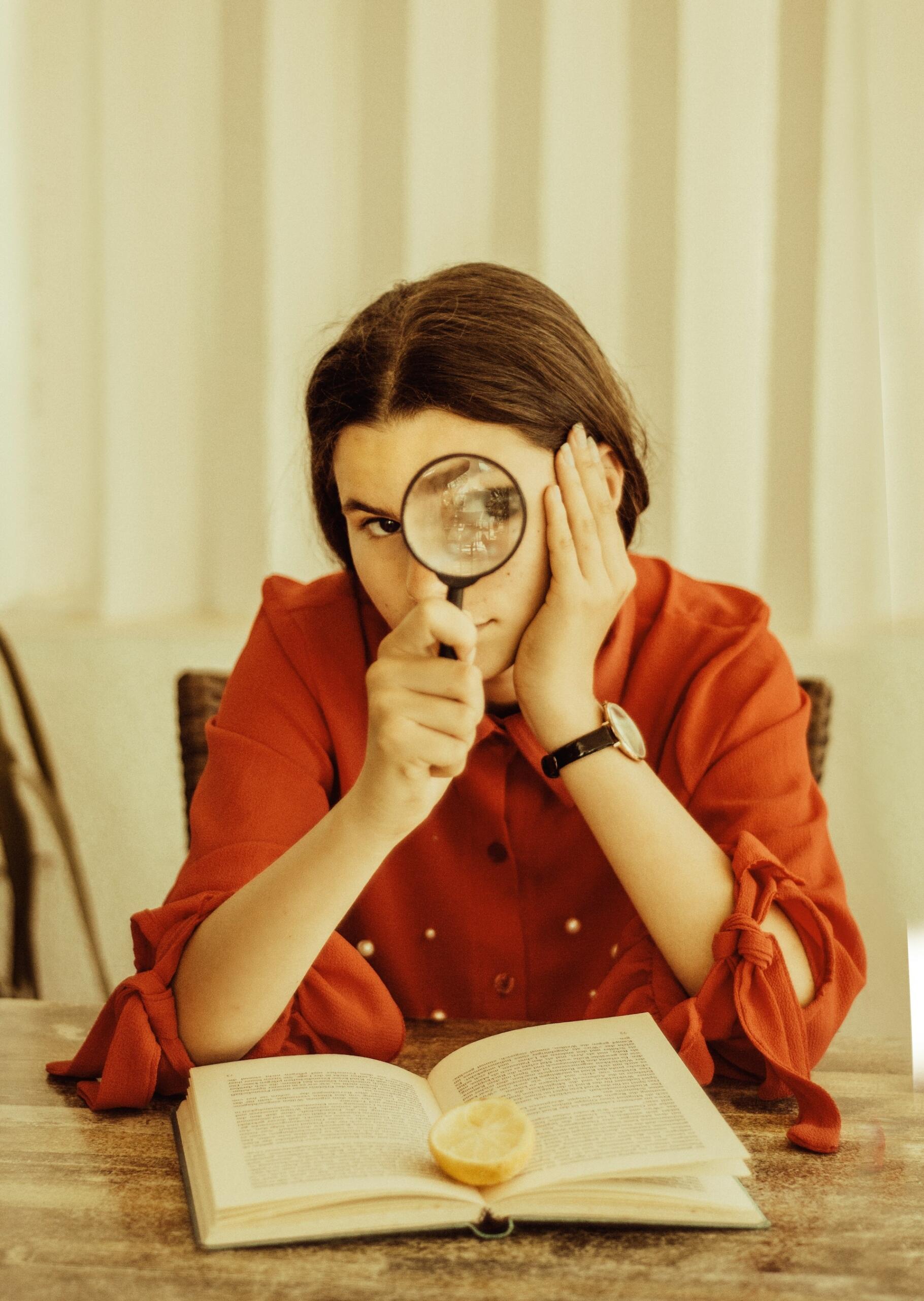 Una persona con una camiseta roja observa a través de una lupa un libro abierto en el que hay un limón, transmitiendo curiosidad y concentración en una habitación con una iluminación tenue.