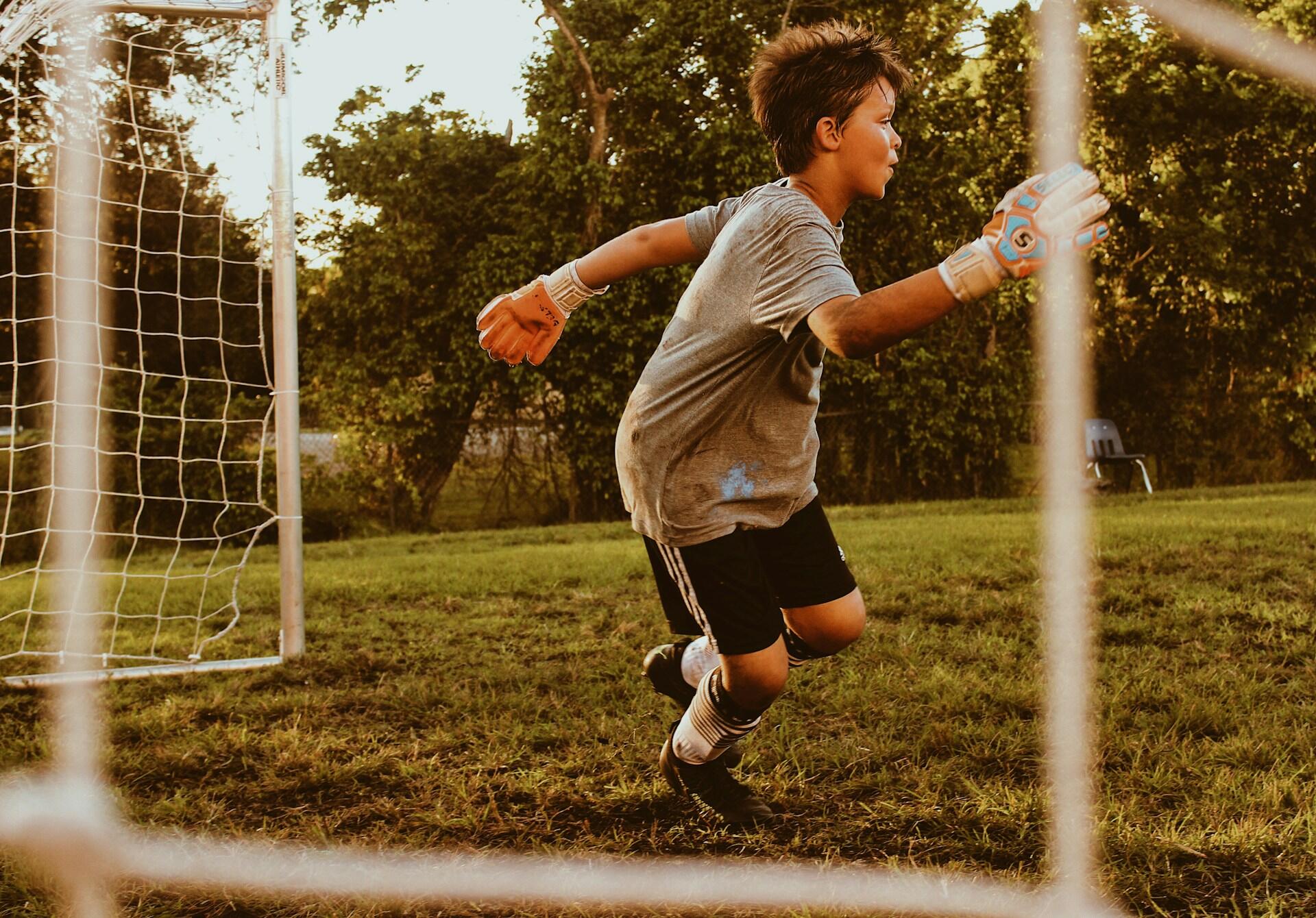 Un joven portero de fútbol vestido de gris y con guantes naranjas se concentra intensamente mientras se mueve con energía cerca del poste de la portería en un campo frondoso bajo la luz dorada del sol.