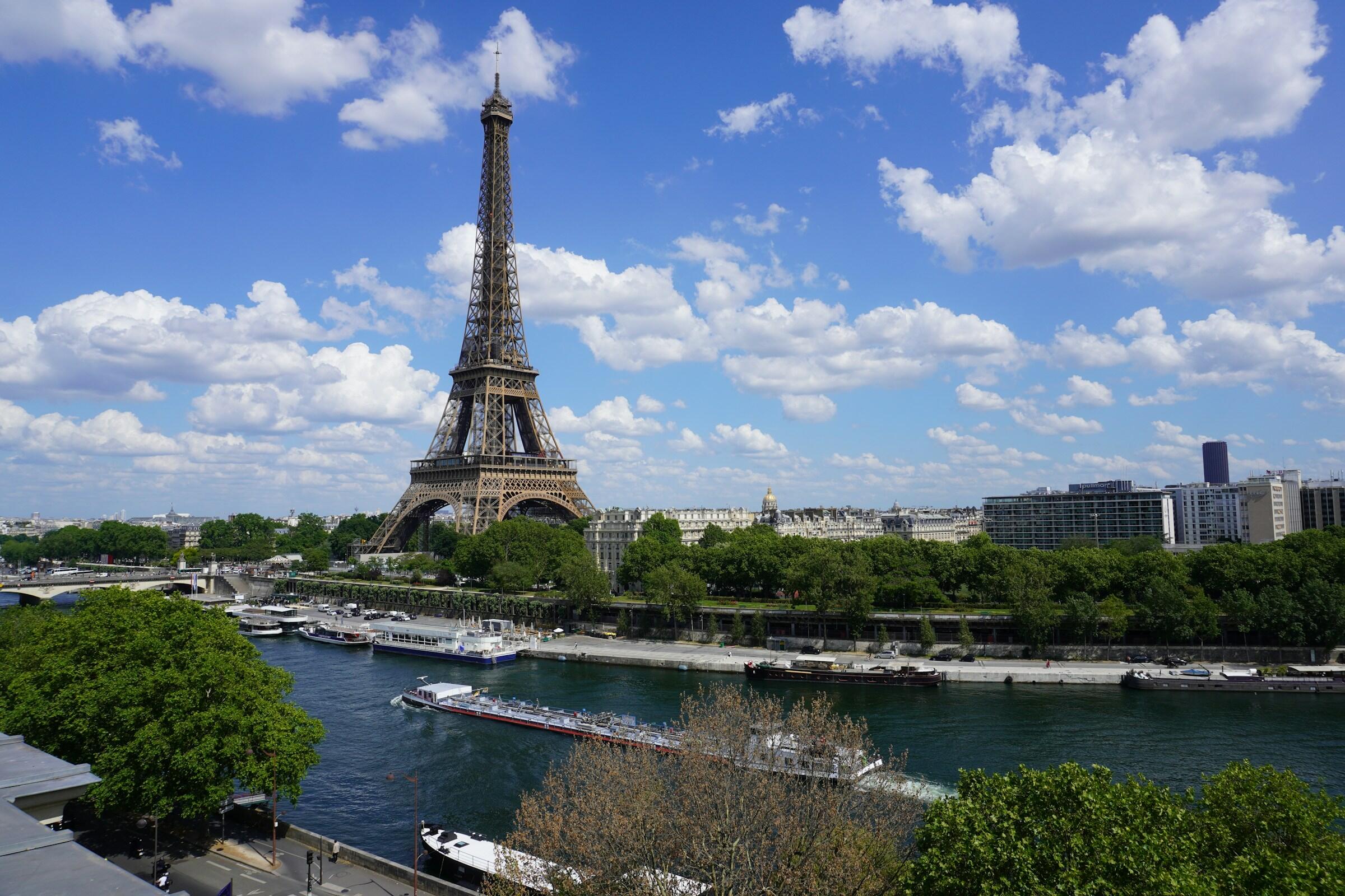 Vista de la Torre Eiffel entre exuberante vegetación, el río Sena y un pintoresco cielo lleno de esponjosas nubes.
