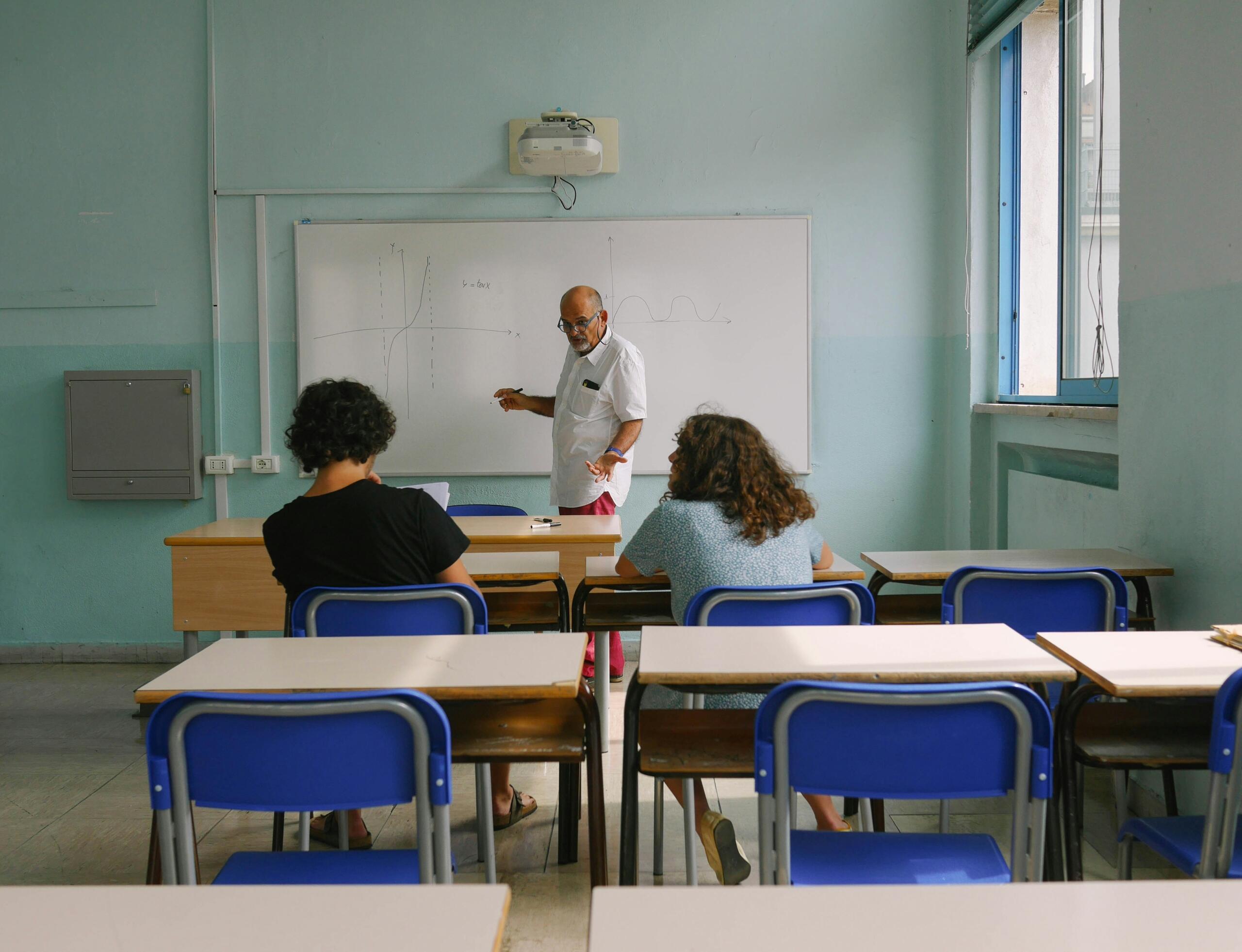 Un profesor junto a una pizarra explica conceptos a dos alumnos sentados en pupitres en un aula azul claro.