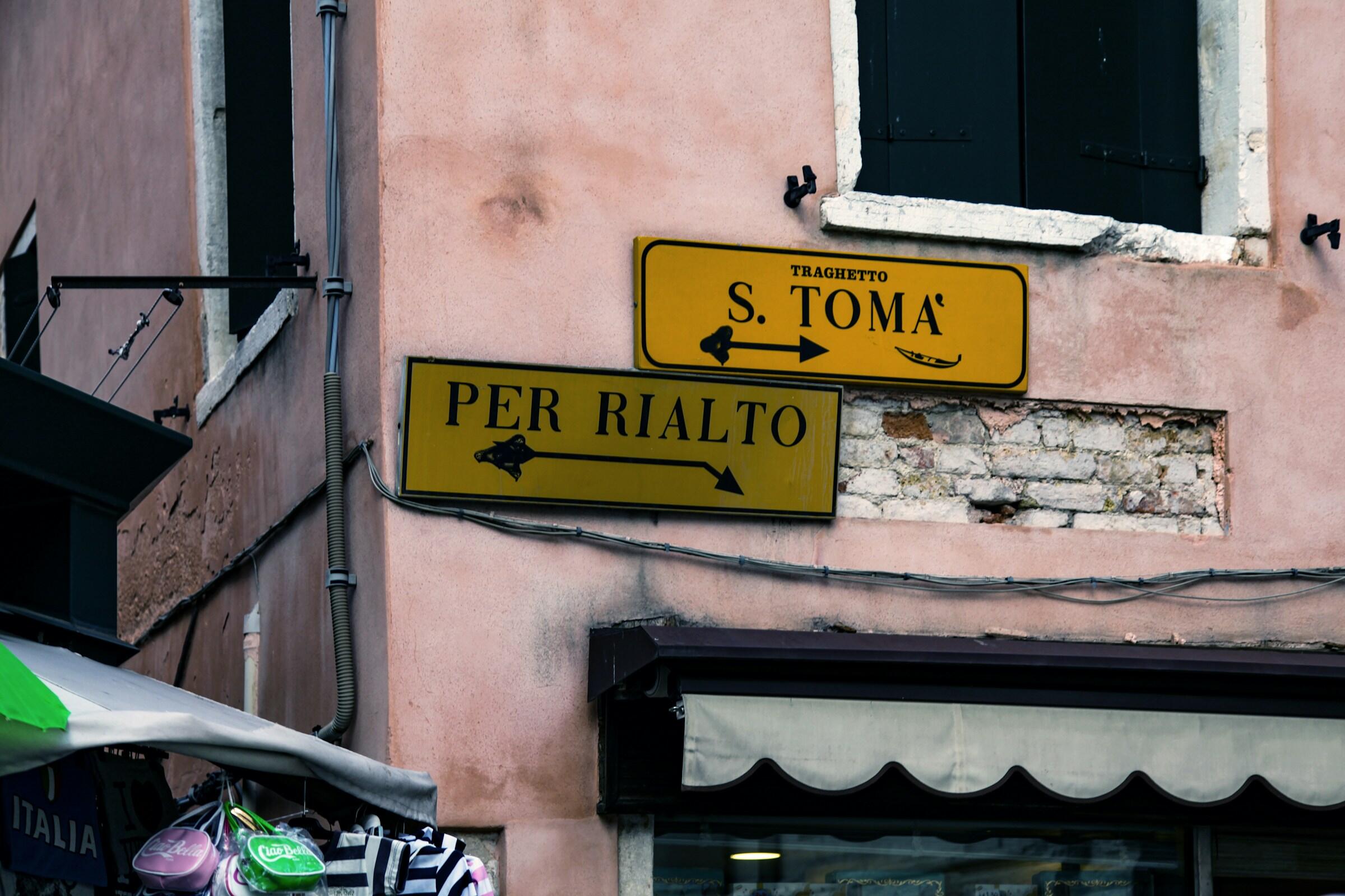 Dos señales de dirección amarillas en la pared de un edificio rosa, indicando caminos hacia «S. Toma» y «Per Rialto».