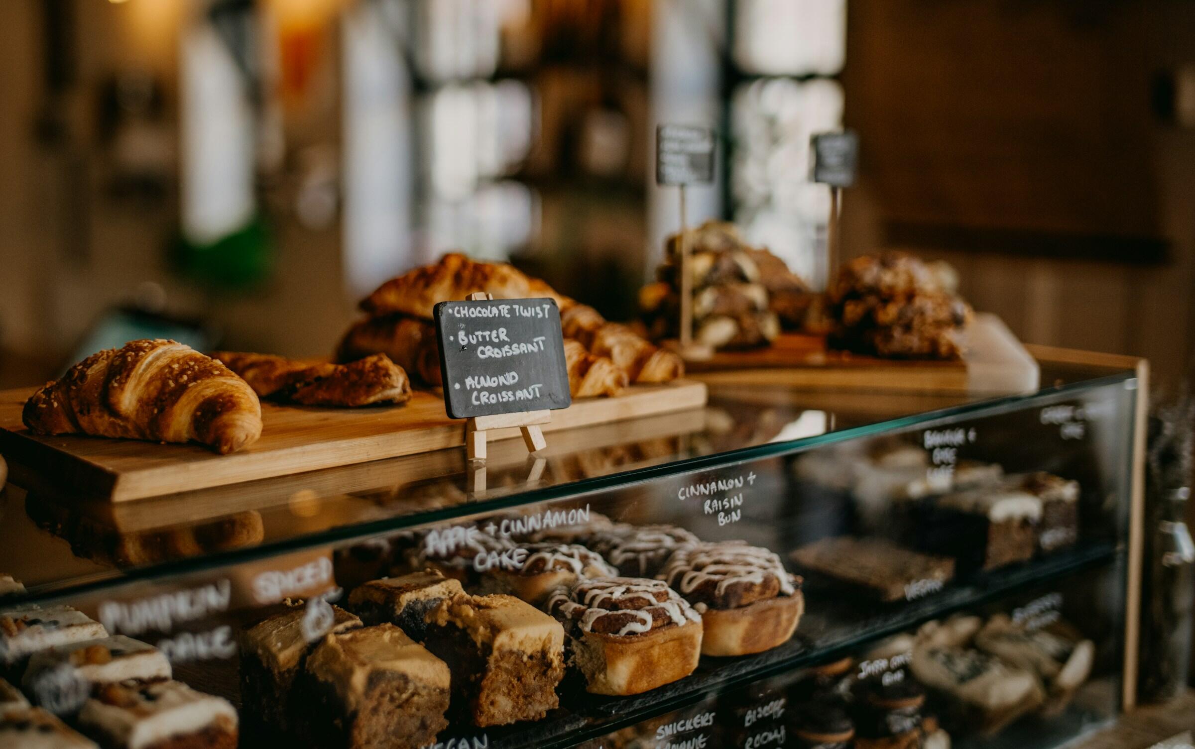 Una vitrina llena de bollería variada, incluidos cruasanes y pasteles, con rótulos etiquetados para cada artículo. Ambiente cálido y acogedor.
