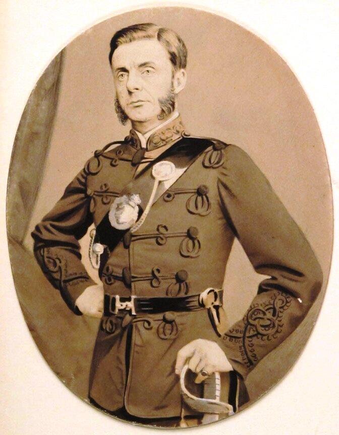 Retrato en sepia de un oficial militar de aspecto severo y con barba, vestido con un uniforme condecorado, empuñando una espada y de pie junto a una bandera cubierta con un paño