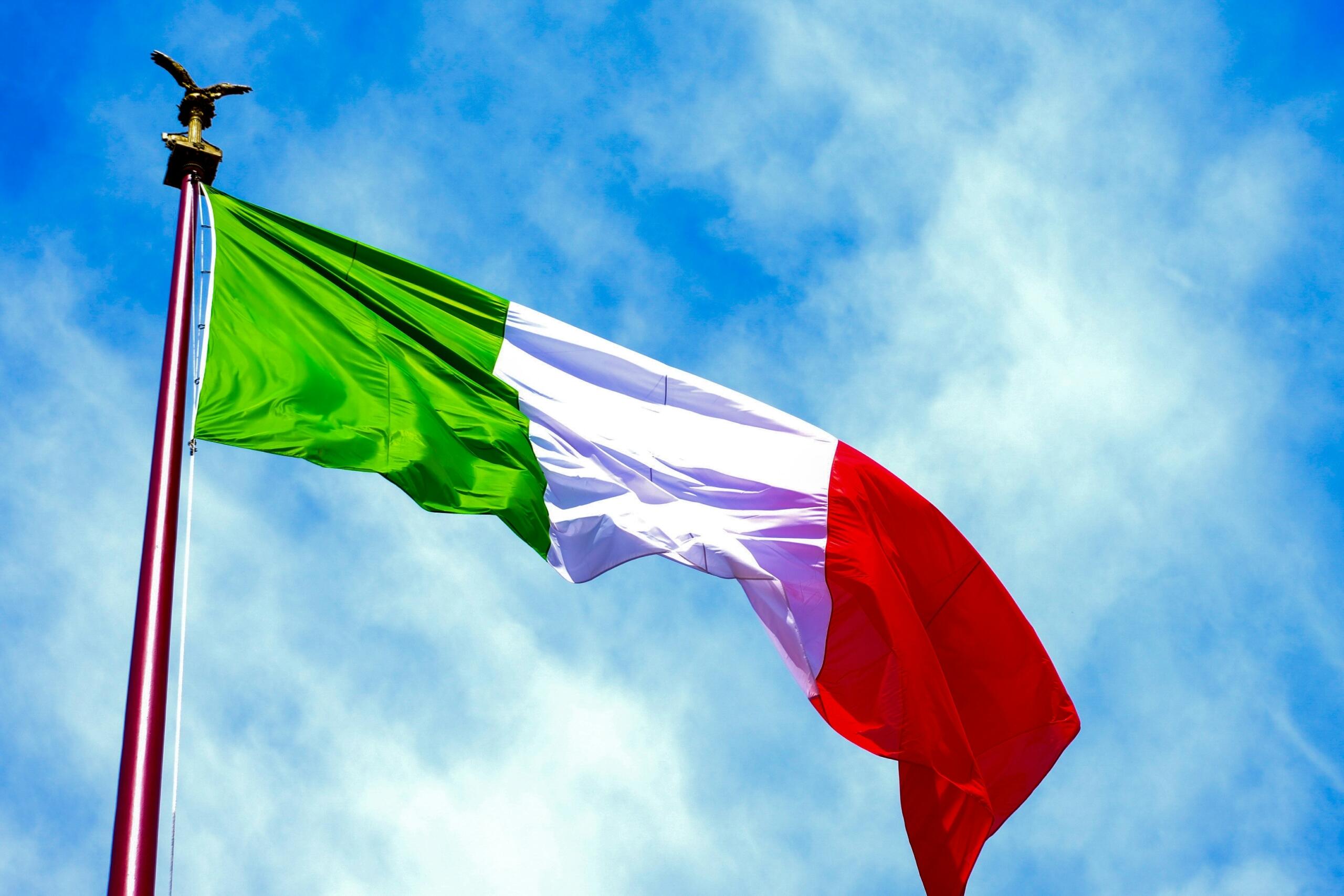 La bandera italiana ondeando majestuosamente sobre un mástil rojo, con un águila dorada posada en lo alto, contra un cielo azul brillante.