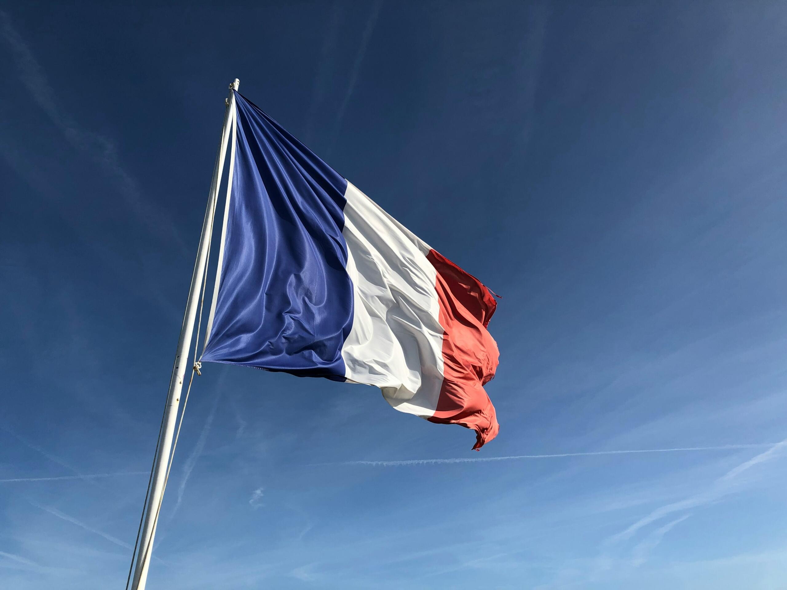 Bandera de Francia ondeando contra un cielo azul despejado, mostrando sus franjas verticales azules, blancas y rojas.