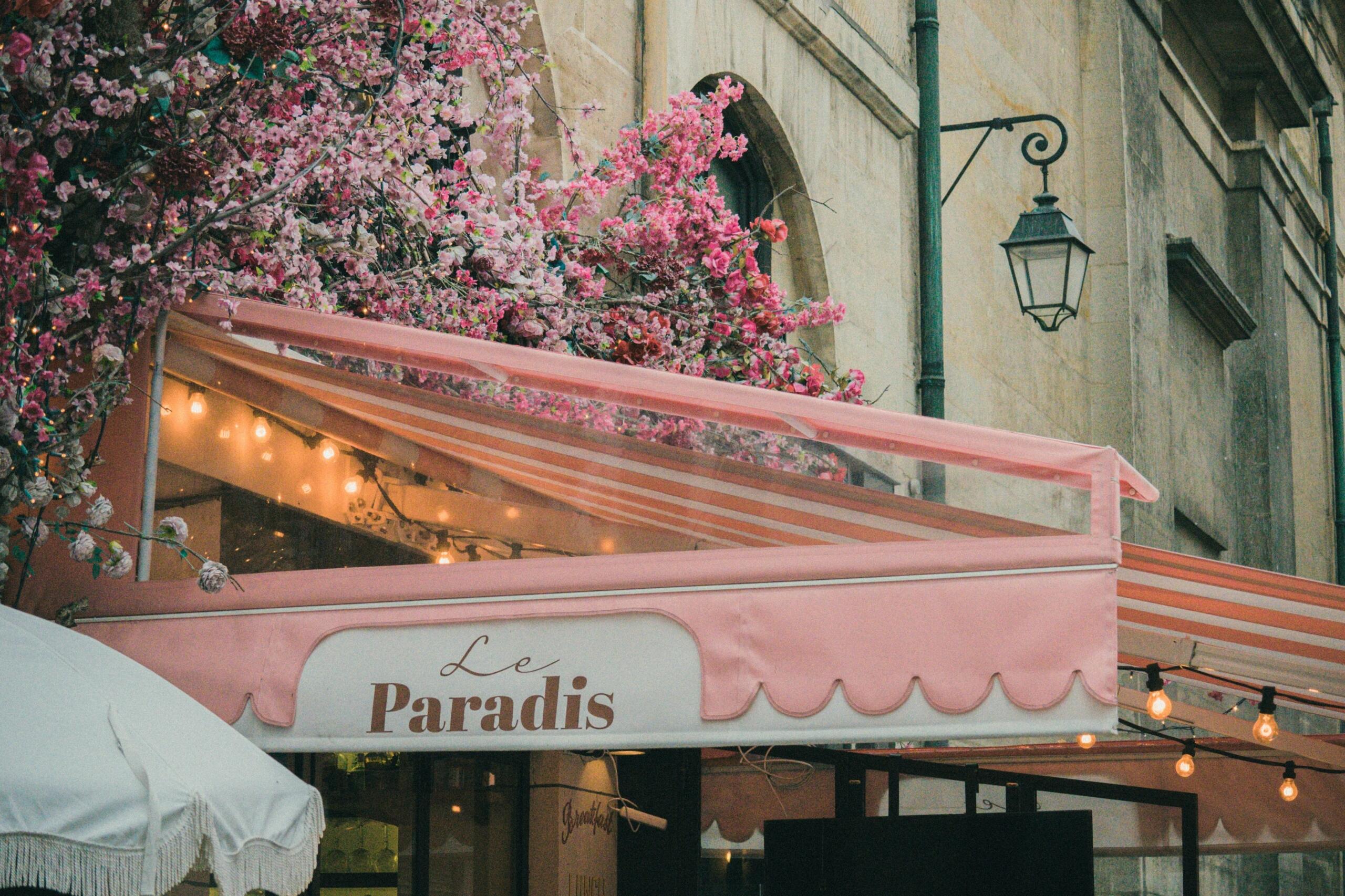 Un encantador café llamado «Le Paradis», adornado con flores y luces rosas, bajo un toldo ruborizado con el telón de fondo de un edificio histórico.