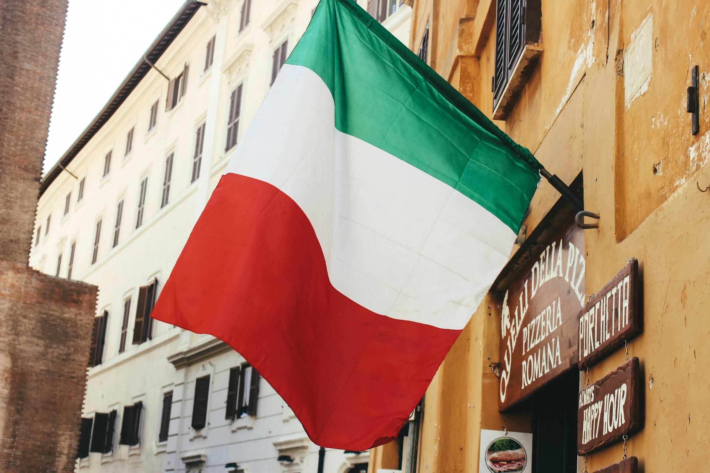 Bandera italiana ondeando frente a una pizzería rústica, con una cálida pared amarilla y carteles de madera que aluden a la cocina tradicional romana.