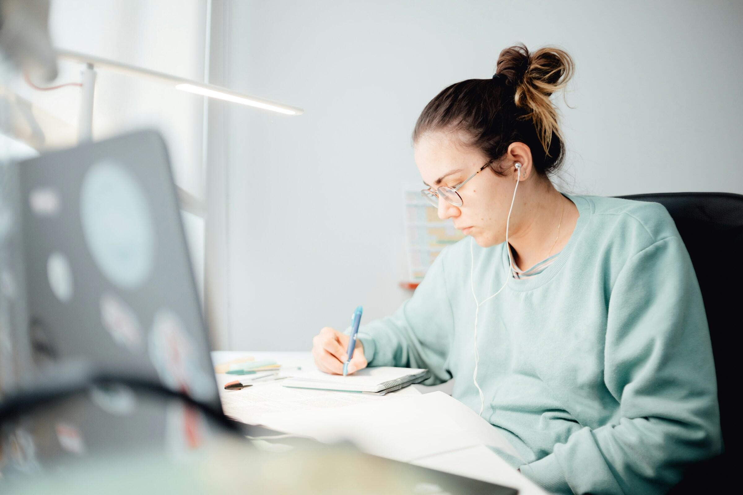 Una persona sentada en un escritorio, con una sudadera ligera y auriculares, escribiendo notas en un papel con un bolígrafo, rodeada de libros y un ordenador portátil.