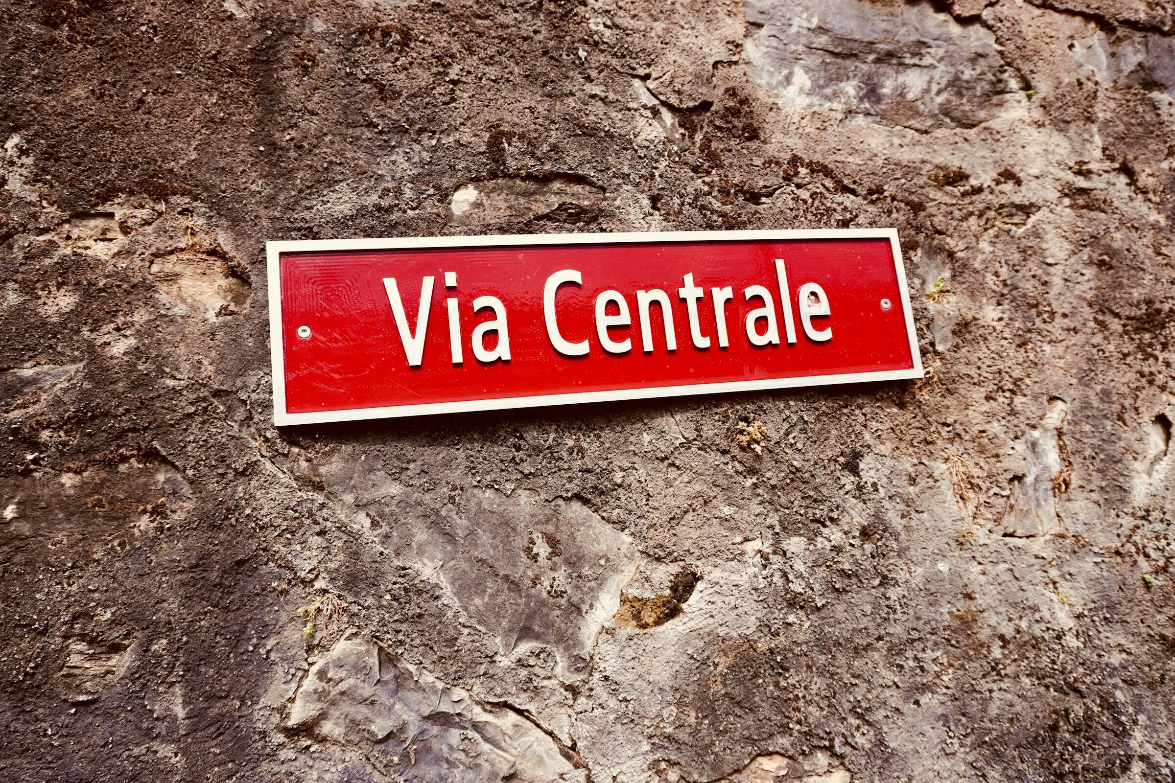 Un letrero rojo en el que se lee «Via Centrale» montado sobre un muro de piedra con textura.