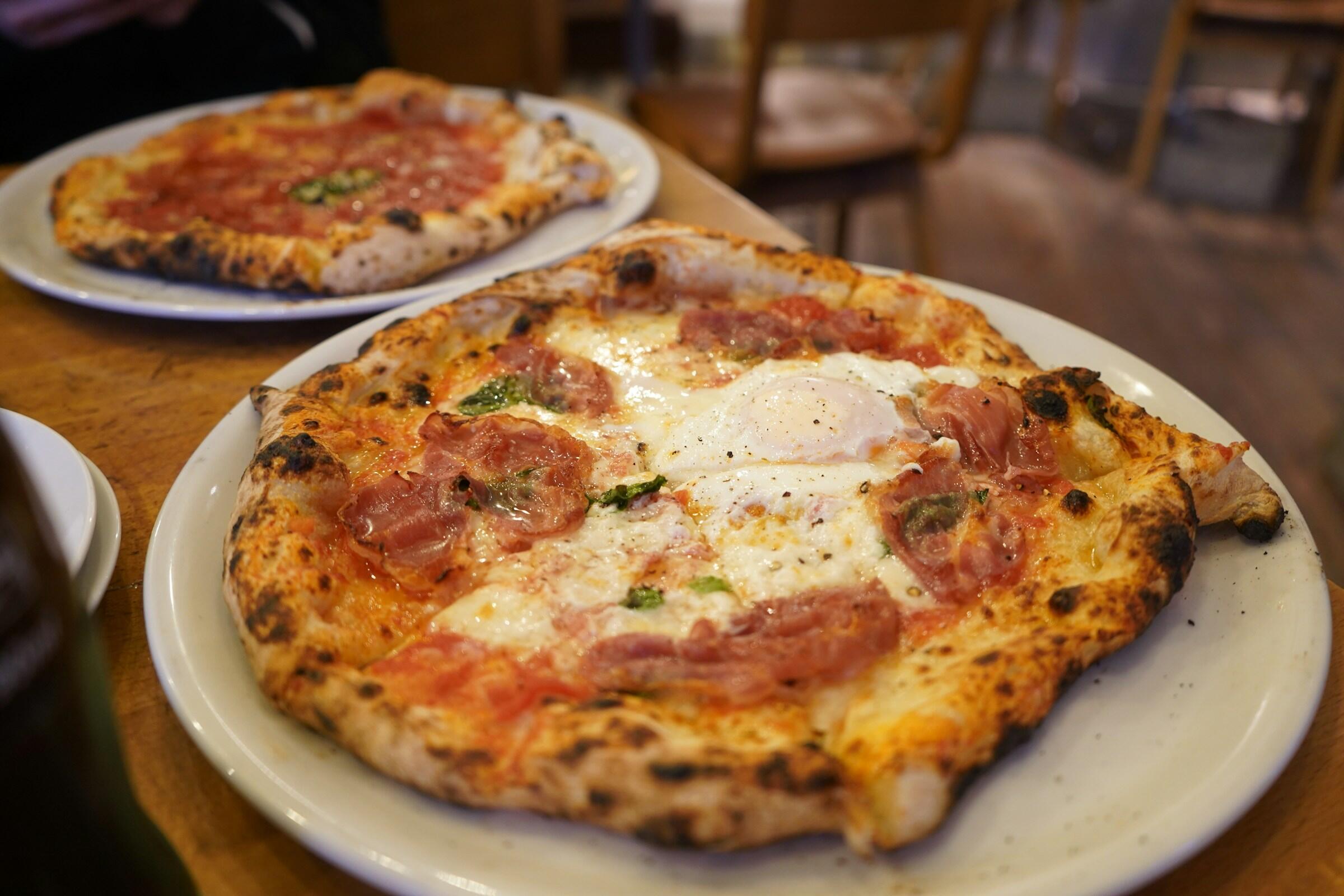 Primer plano de una pizza recién horneada con jamón york, huevo cremoso y hierbas verdes, servida en un plato blanco.