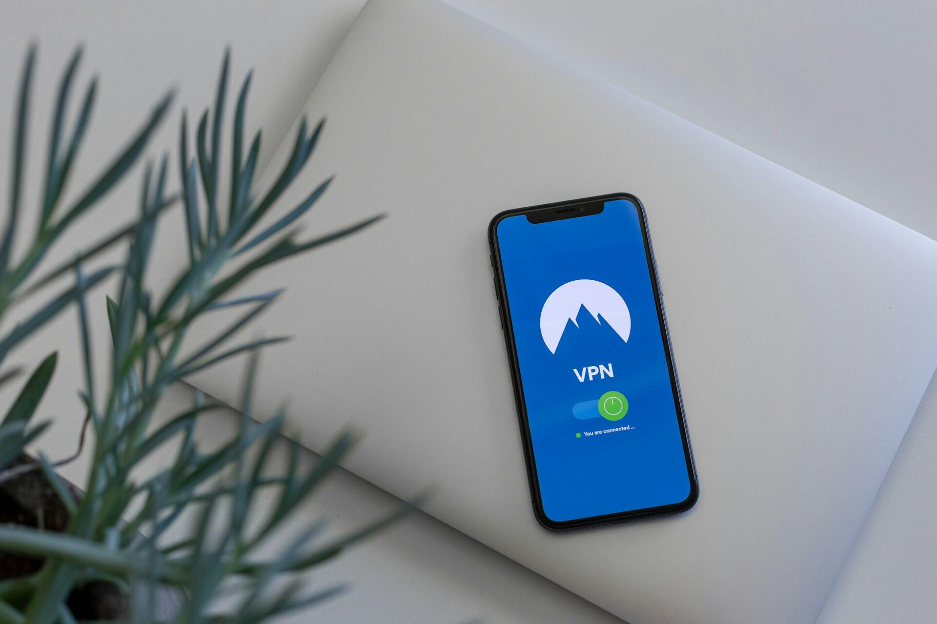 Un smartphone en el que se ve una aplicación de VPN con una pantalla azul y un logotipo de una montaña, apoyado sobre un portátil cerrado. Se ve parcialmente una planta, lo que crea un ambiente tecnológico tranquilo.