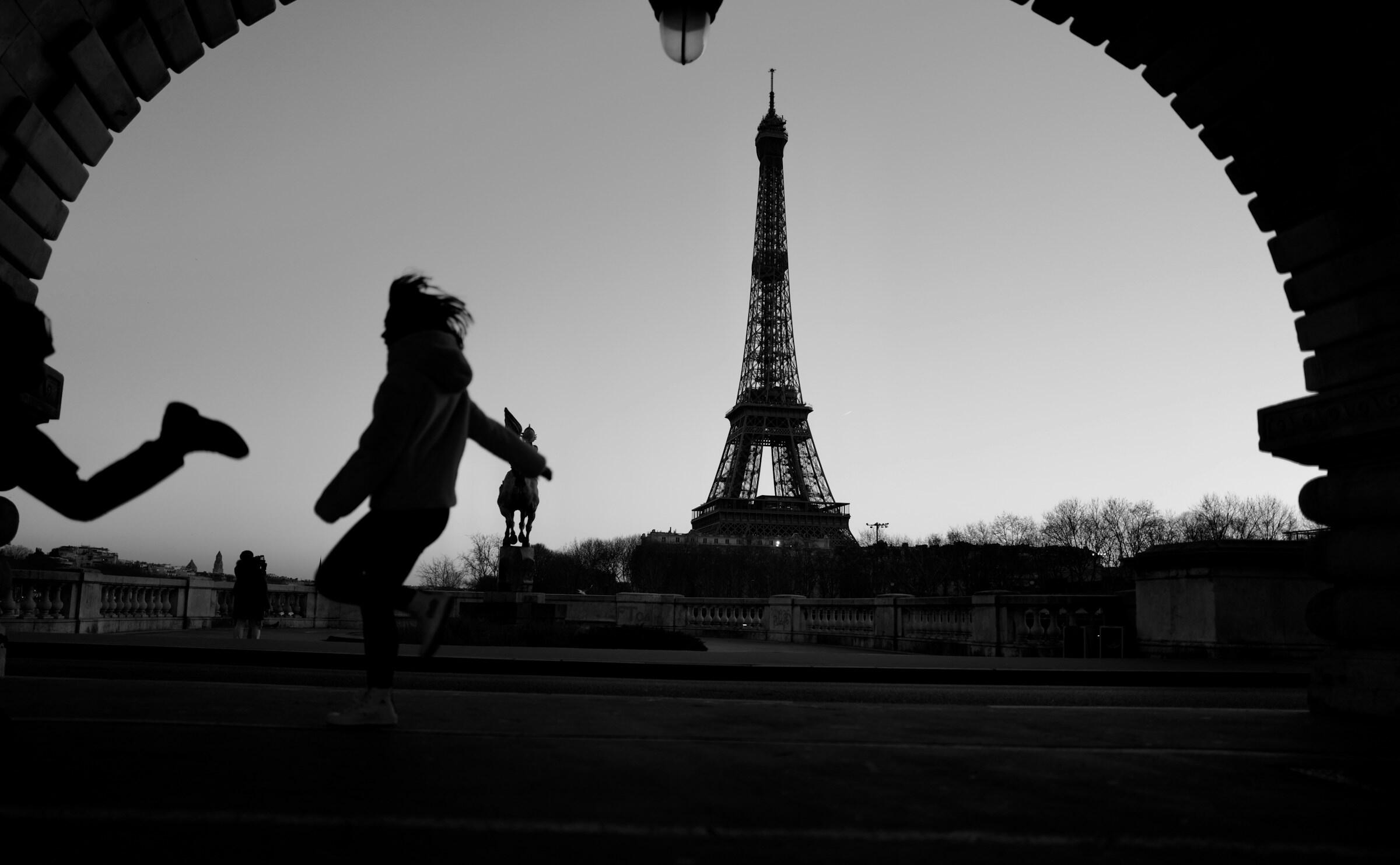 Siluetas de figuras juguetonas frente a la Torre Eiffel al atardecer, enmarcadas por un arco arquitectónico en blanco y negro.