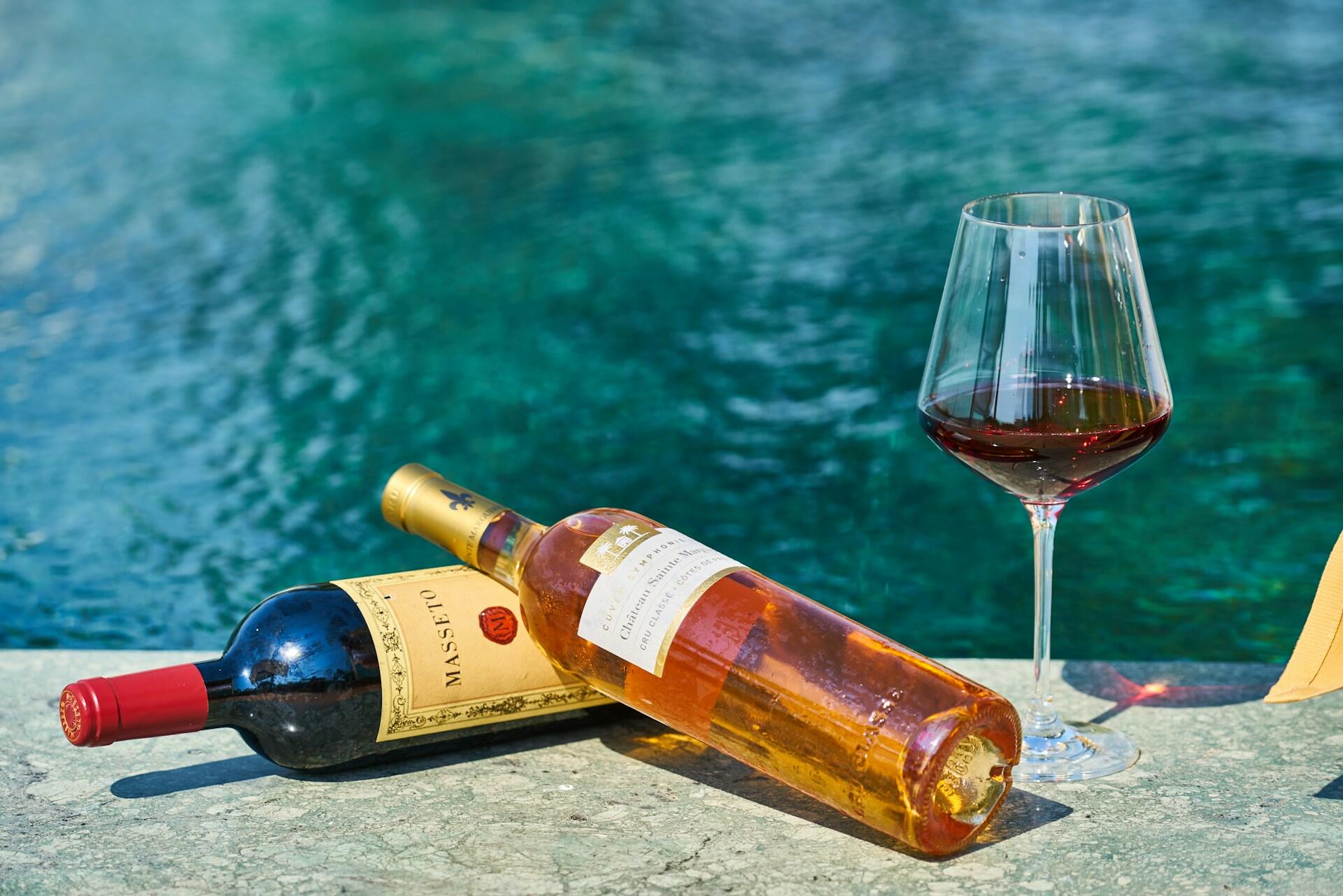 Dos botellas de vino, una tinto y otra rosado, colocadas sobre una superficie de piedra junto a una copa de vino tinto, con un fondo de agua turquesa.