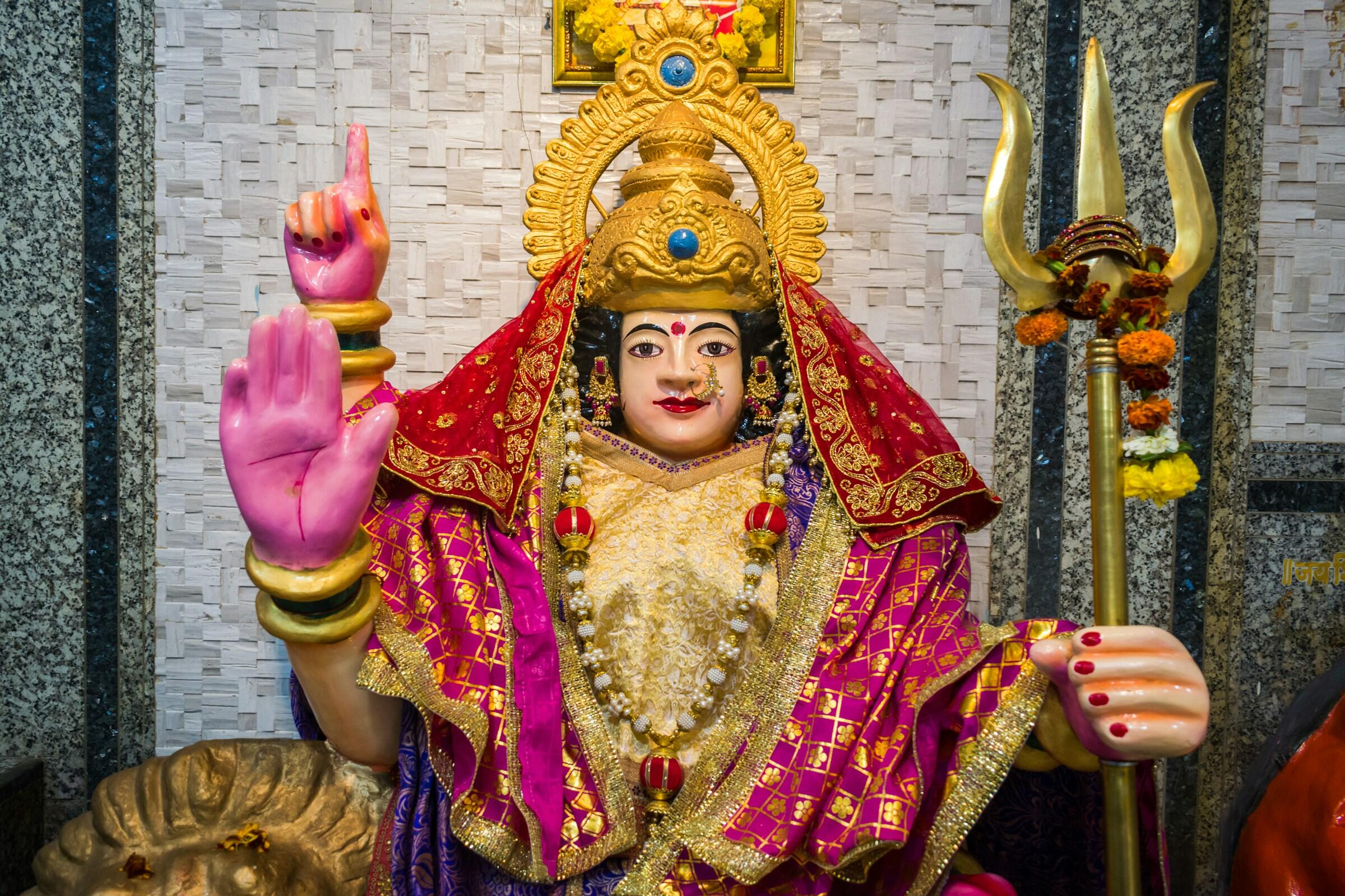 Una colorida estatua adornada con intrincadas joyas y un vibrante atuendo rojo y dorado, que sostiene un tridente y muestra un gesto con la mano.