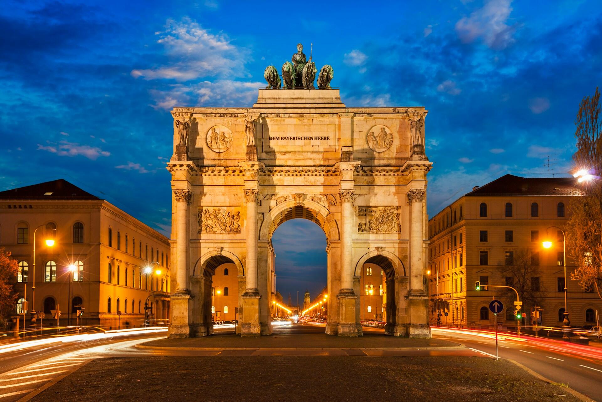 Arco del Triunfo iluminado en Múnich al atardecer, con estatuas en lo alto y elaboradas tallas, con los rastros de los semáforos en primer plano.