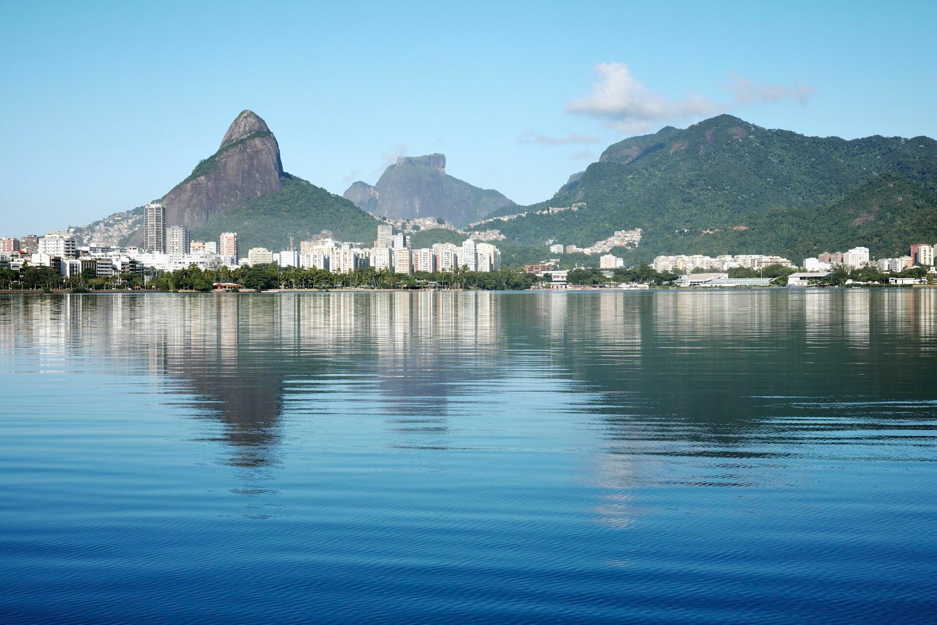 Vista panorámica del horizonte de Río de Janeiro reflejado en aguas tranquilas, con emblemáticas montañas que se elevan al fondo bajo un cielo azul claro.