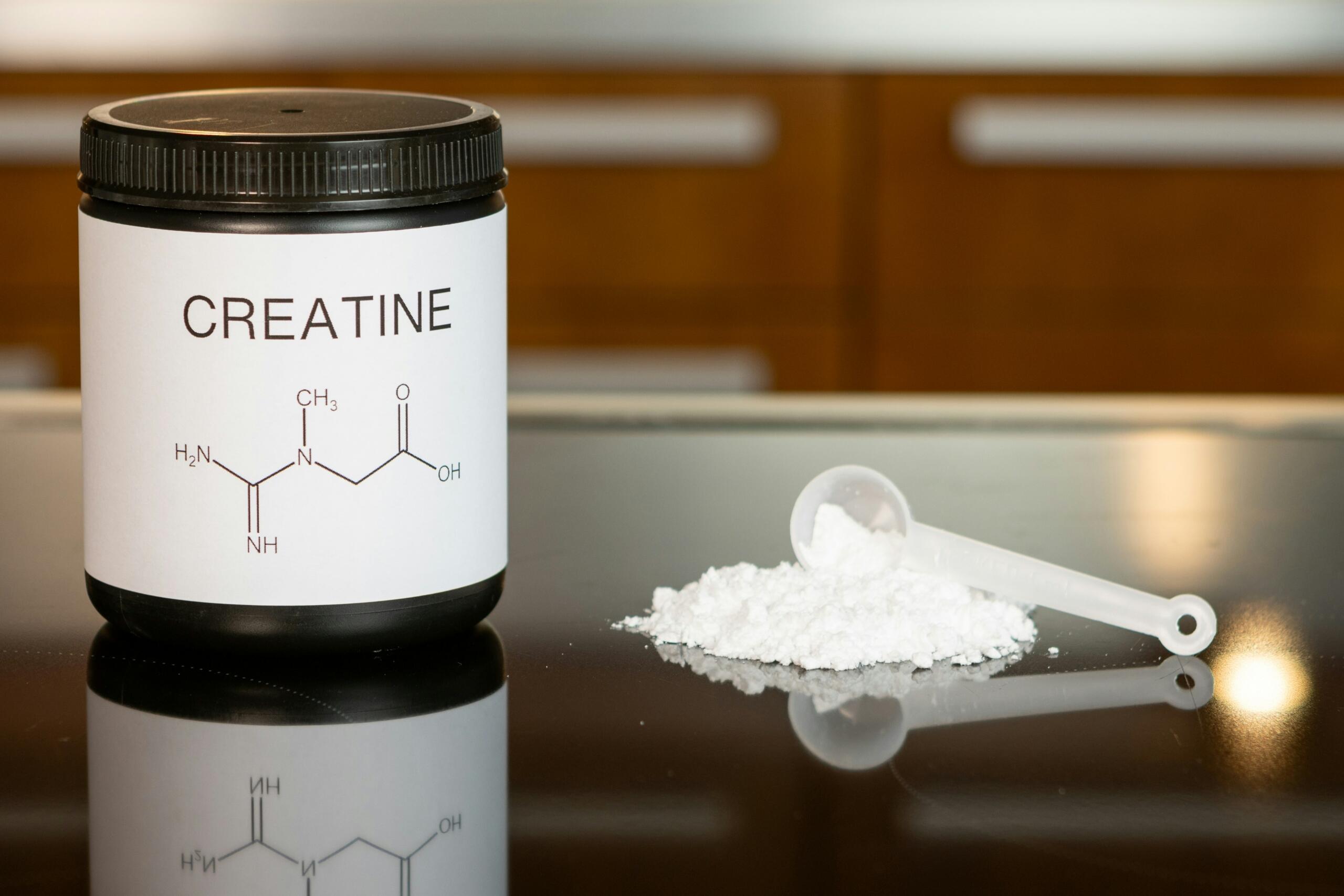 Un tarro negro con la etiqueta «Creatina» y una estructura molecular, junto a una cuchara y un montón de polvo blanco sobre una superficie reflectante.