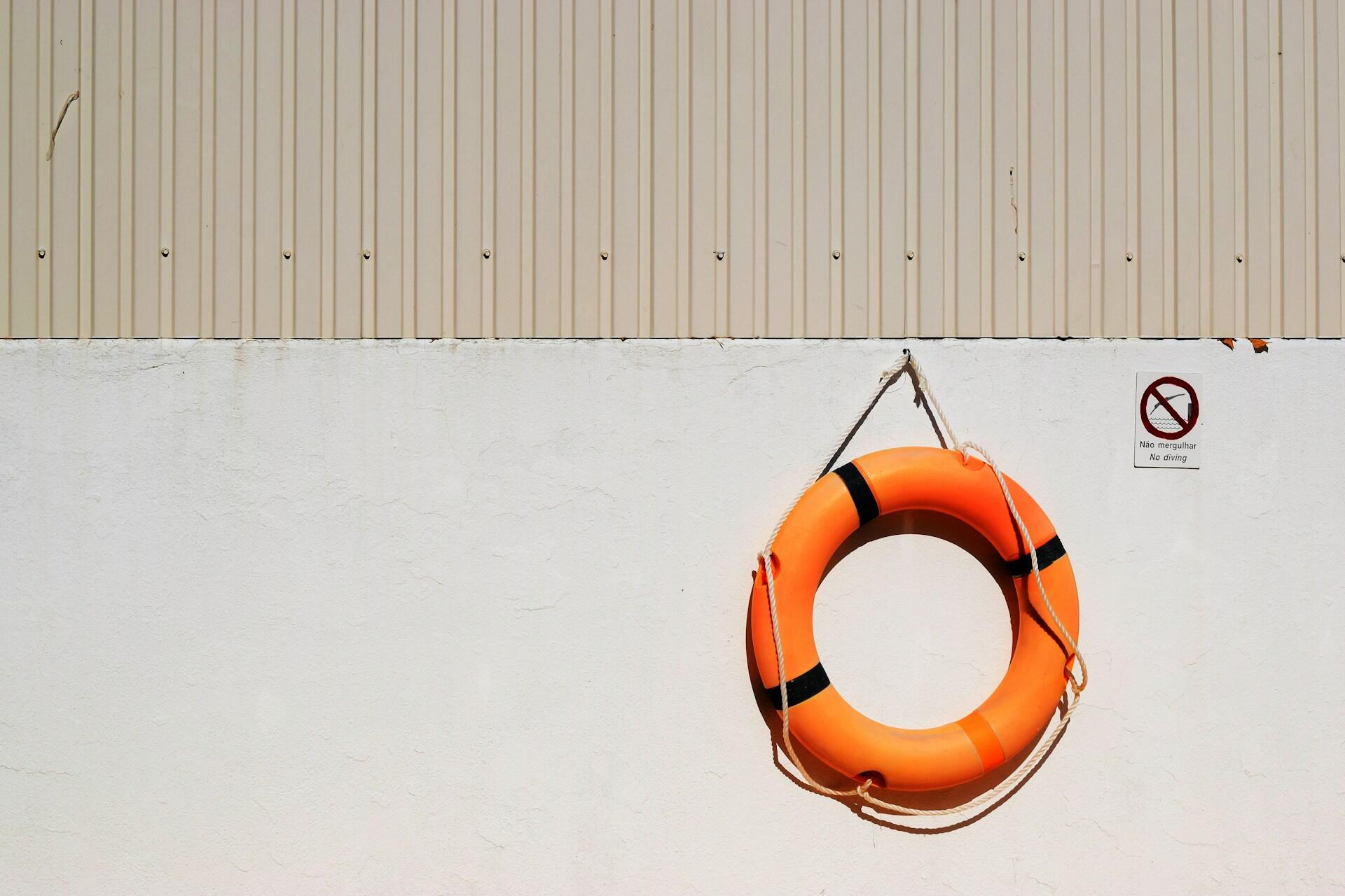 Un salvavidas de color naranja brillante cuelga de una pared blanca, junto a un cartel de «Prohibido bucear», sobre un fondo beige texturizado.