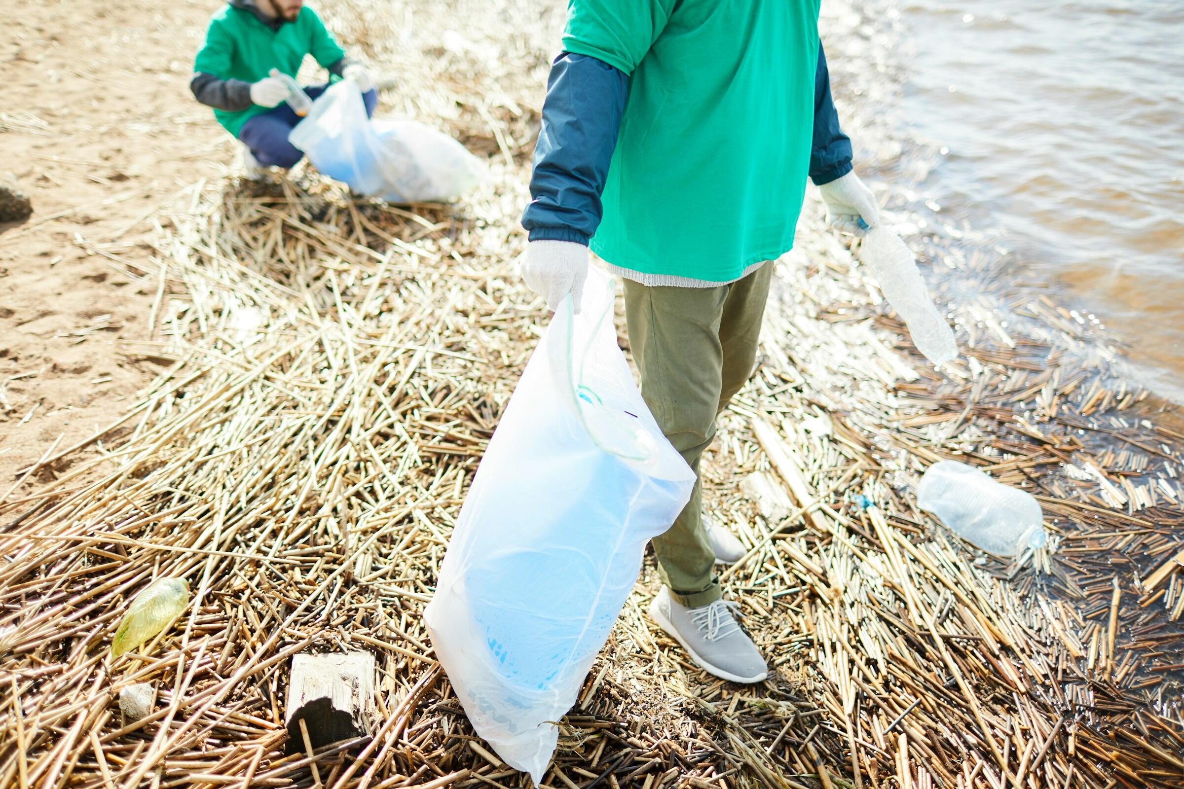 Dos voluntarios con camisas verdes recogen la basura de la playa, llenando bolsas con restos de plástico y materia orgánica a lo largo de la orilla.