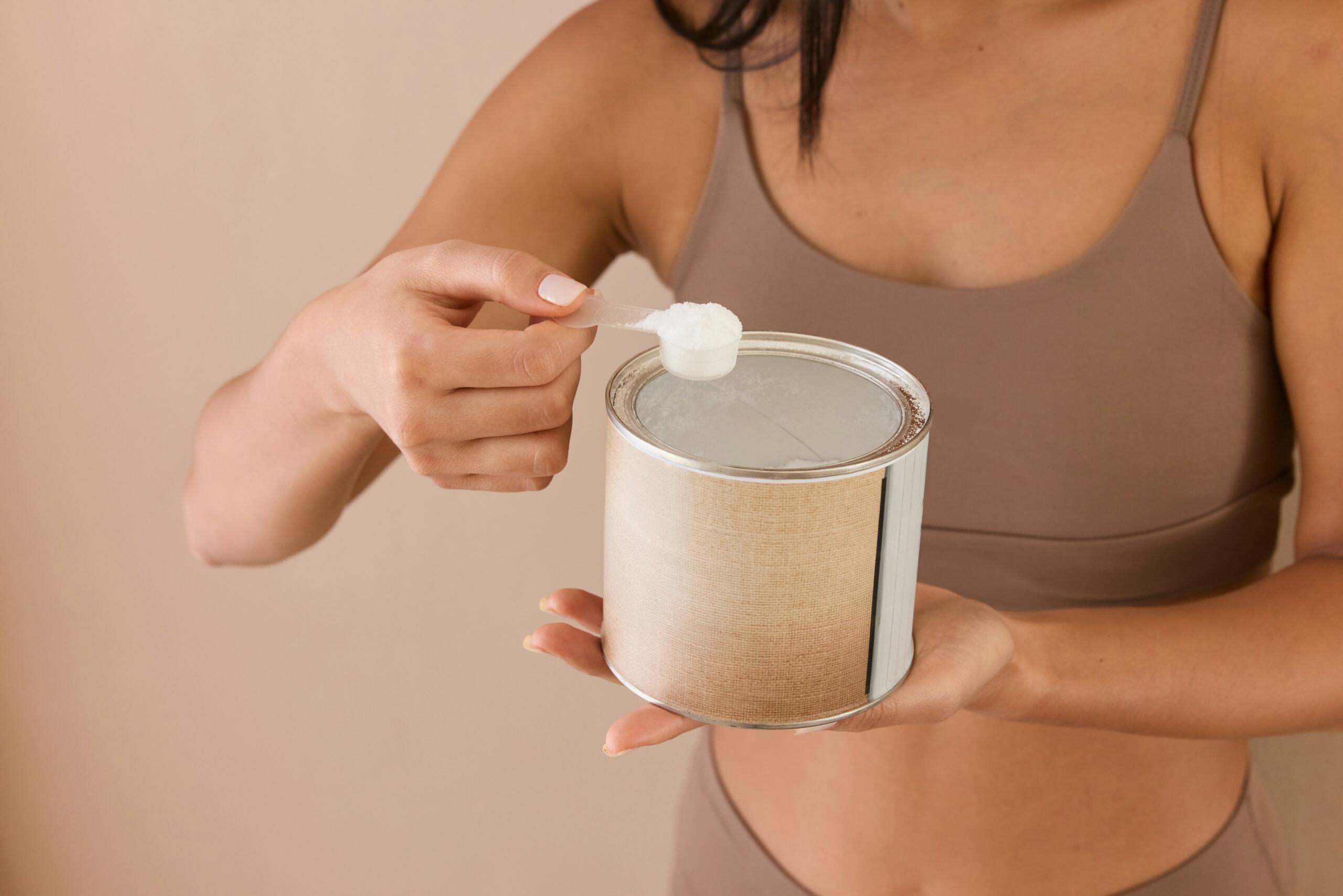Una mujer vestida con ropa deportiva marrón sostiene un bote y saca un polvo blanco con una cuchara dosificadora. Fondo neutro.