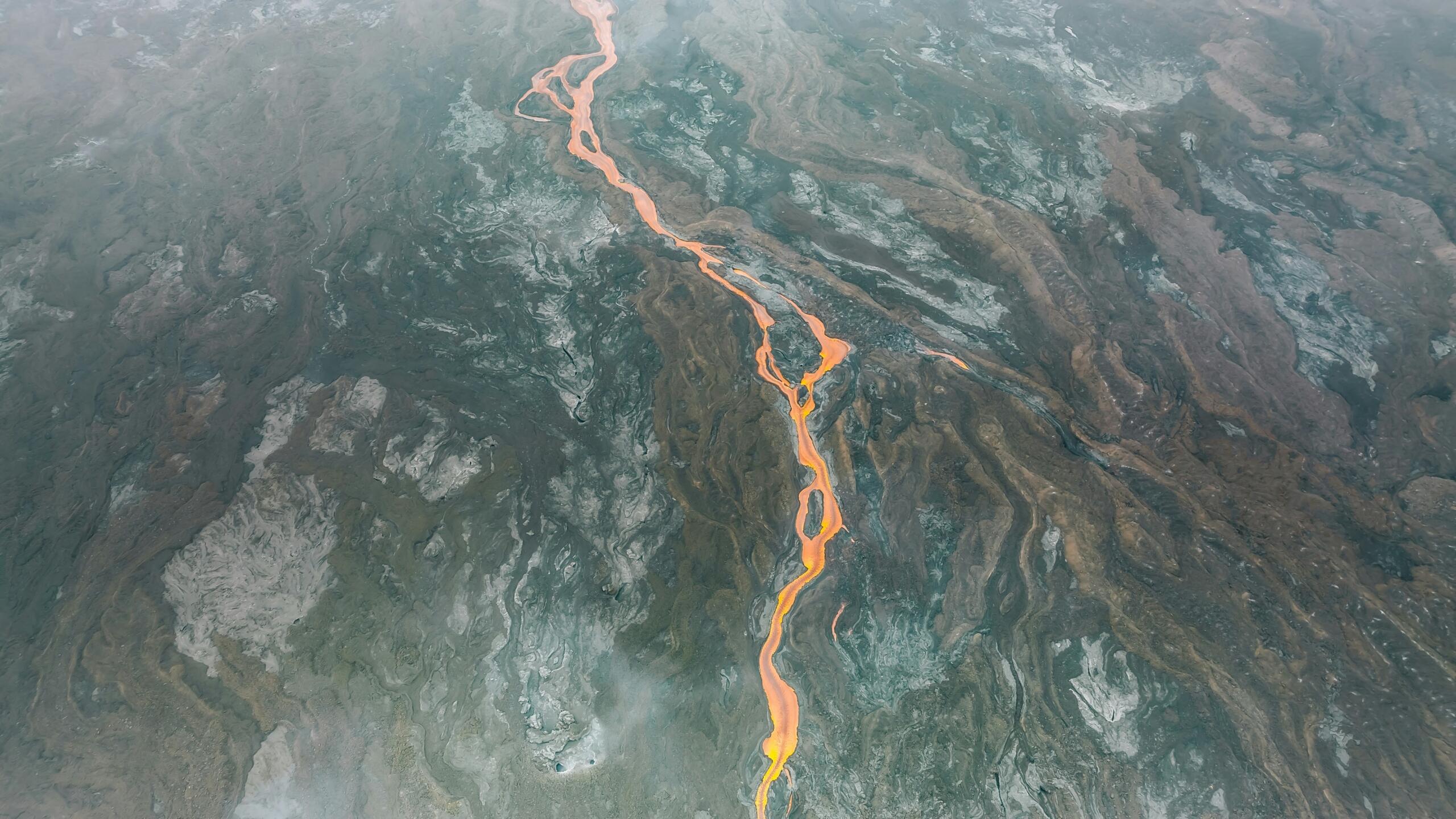 Vista aérea de un vibrante y caudaloso río de lava que serpentea por un terreno volcánico oscuro y texturado, mostrando la actividad geotérmica natural.