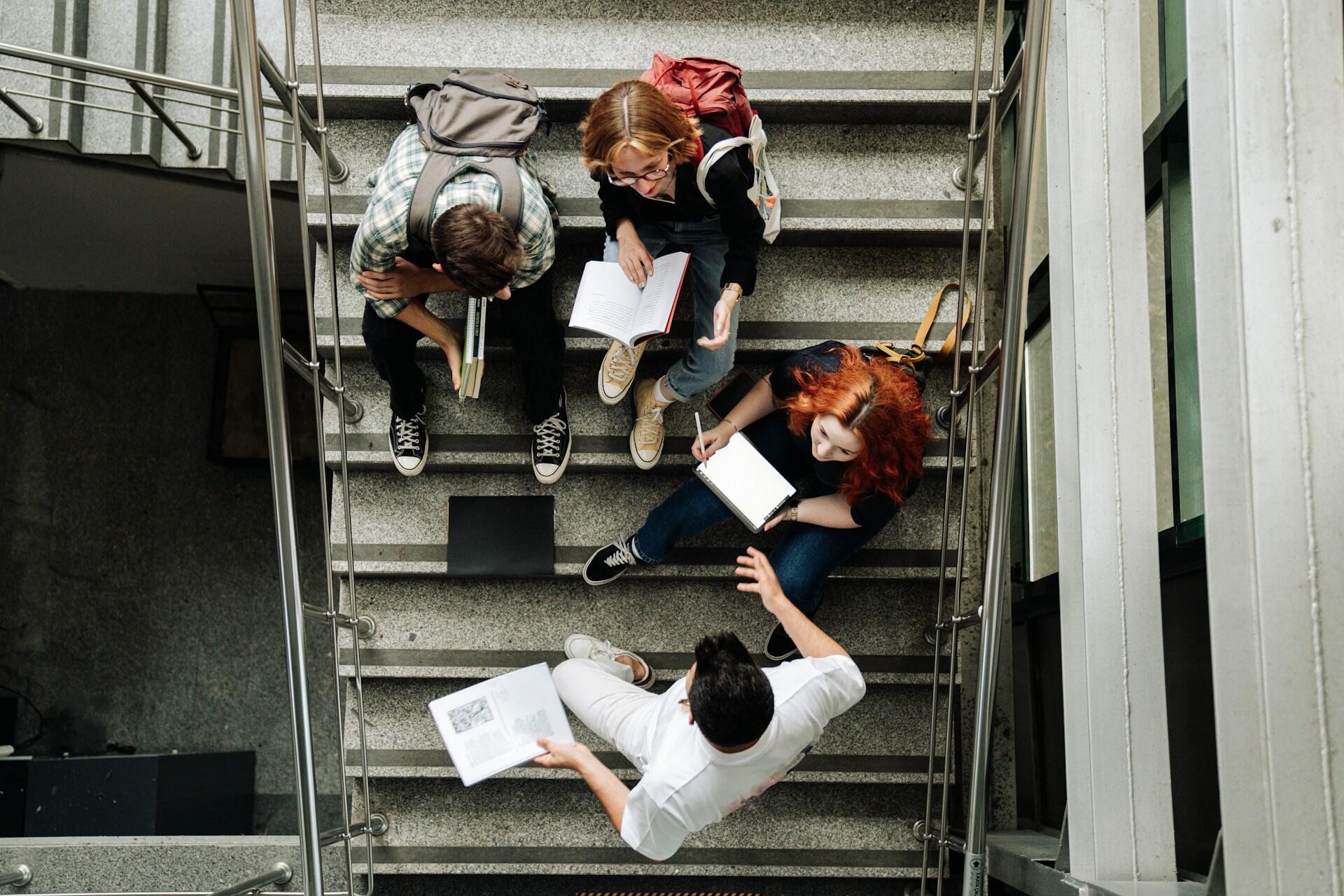 Un grupo de estudiantes estudian juntos en las escaleras, inmersos en libros y apuntes en un entorno de aprendizaje colaborativo.