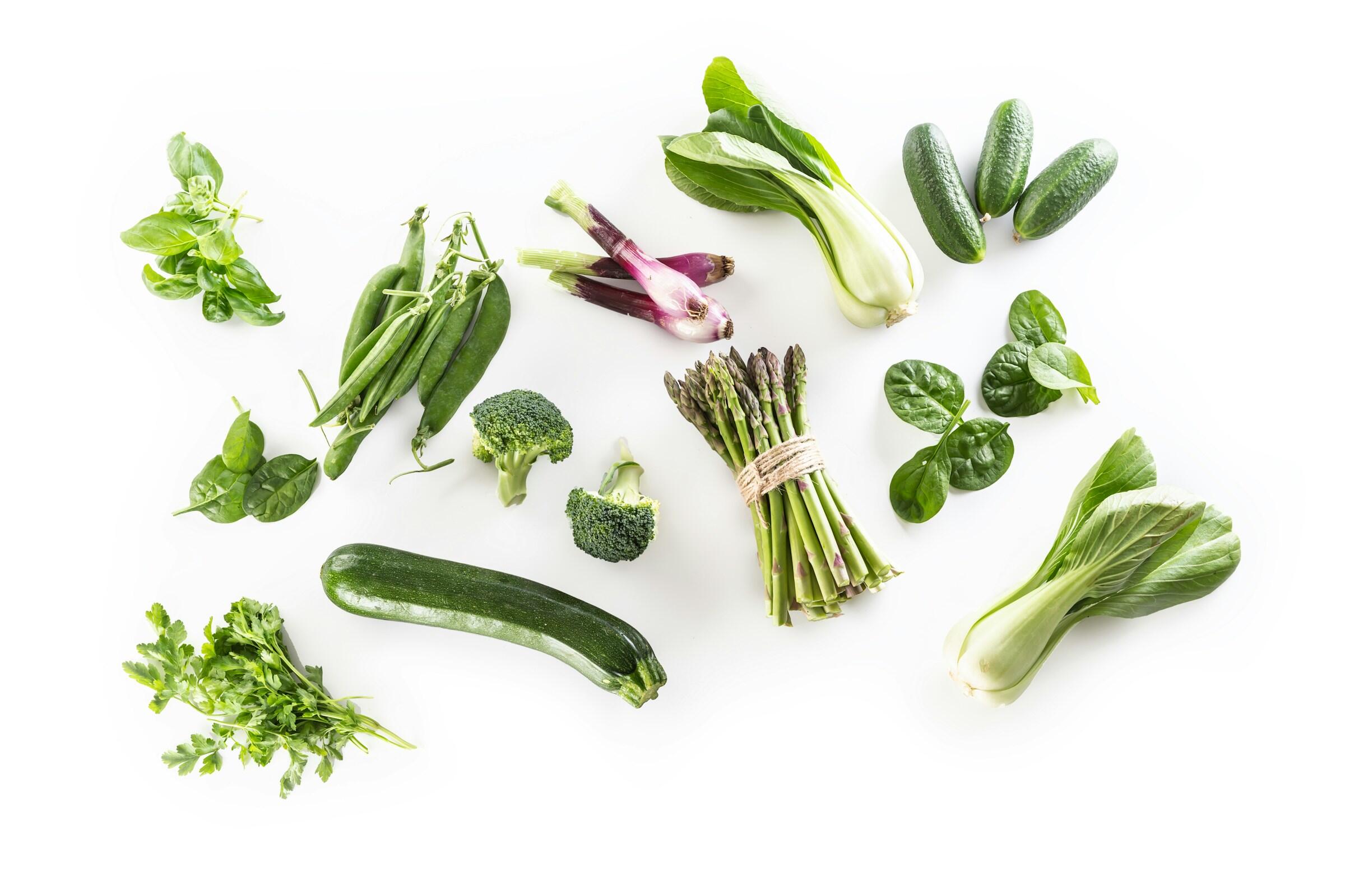 Surtido de verduras verdes frescas, como pepinos, brócoli, espárragos y verduras de hoja verde, dispuestas artísticamente sobre un fondo blanco.