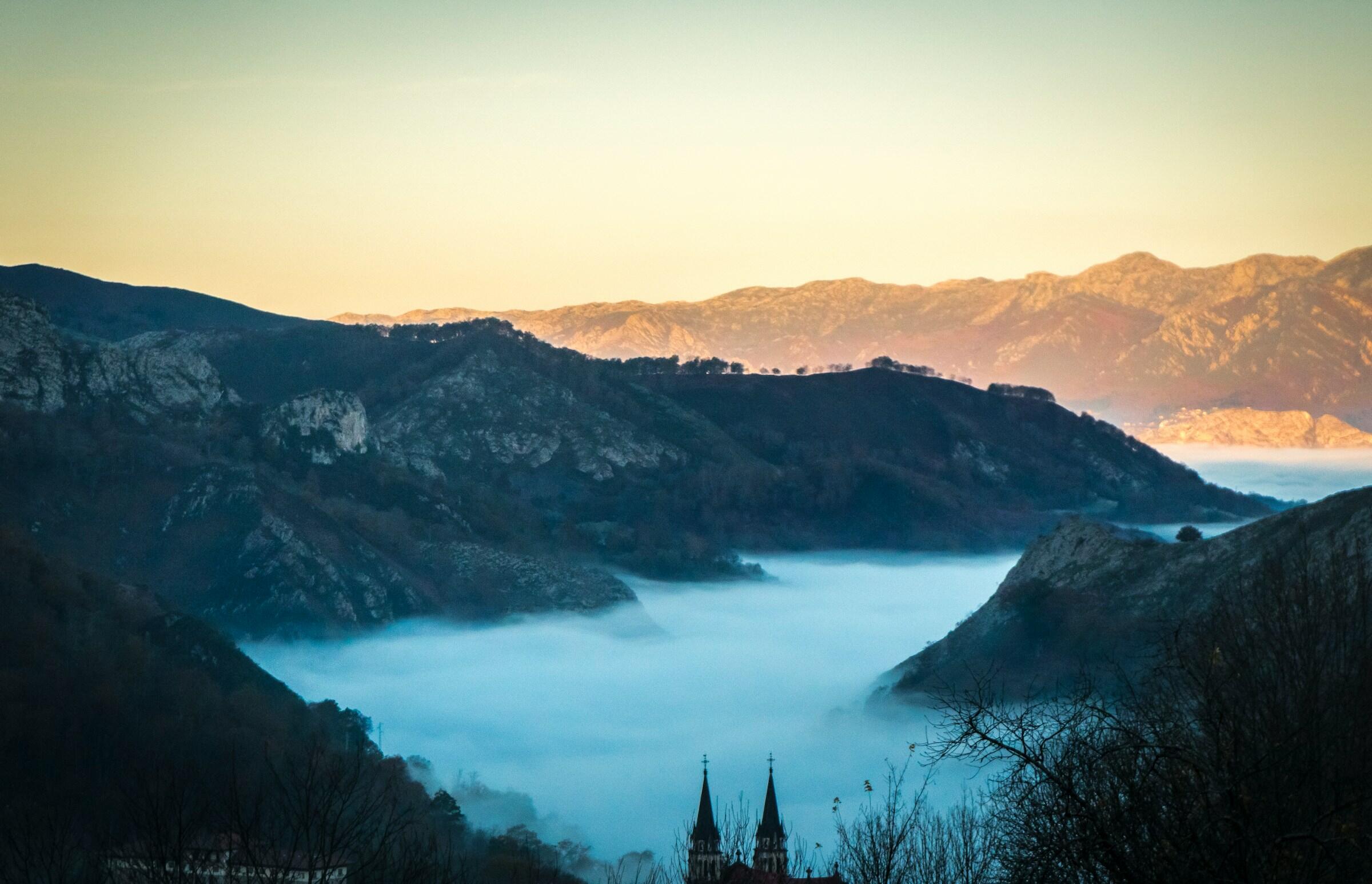 Sereno paisaje de montaña al amanecer, con la niebla ondulando por los valles y el campanario de una iglesia lejana asomando desde abajo en los lagos de covadonga