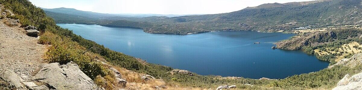 Una vista panorámica del lago de sanabria rodeado de exuberantes colinas verdes bajo un cielo azul despejado, que muestra un terreno accidentado y montañas lejanas.