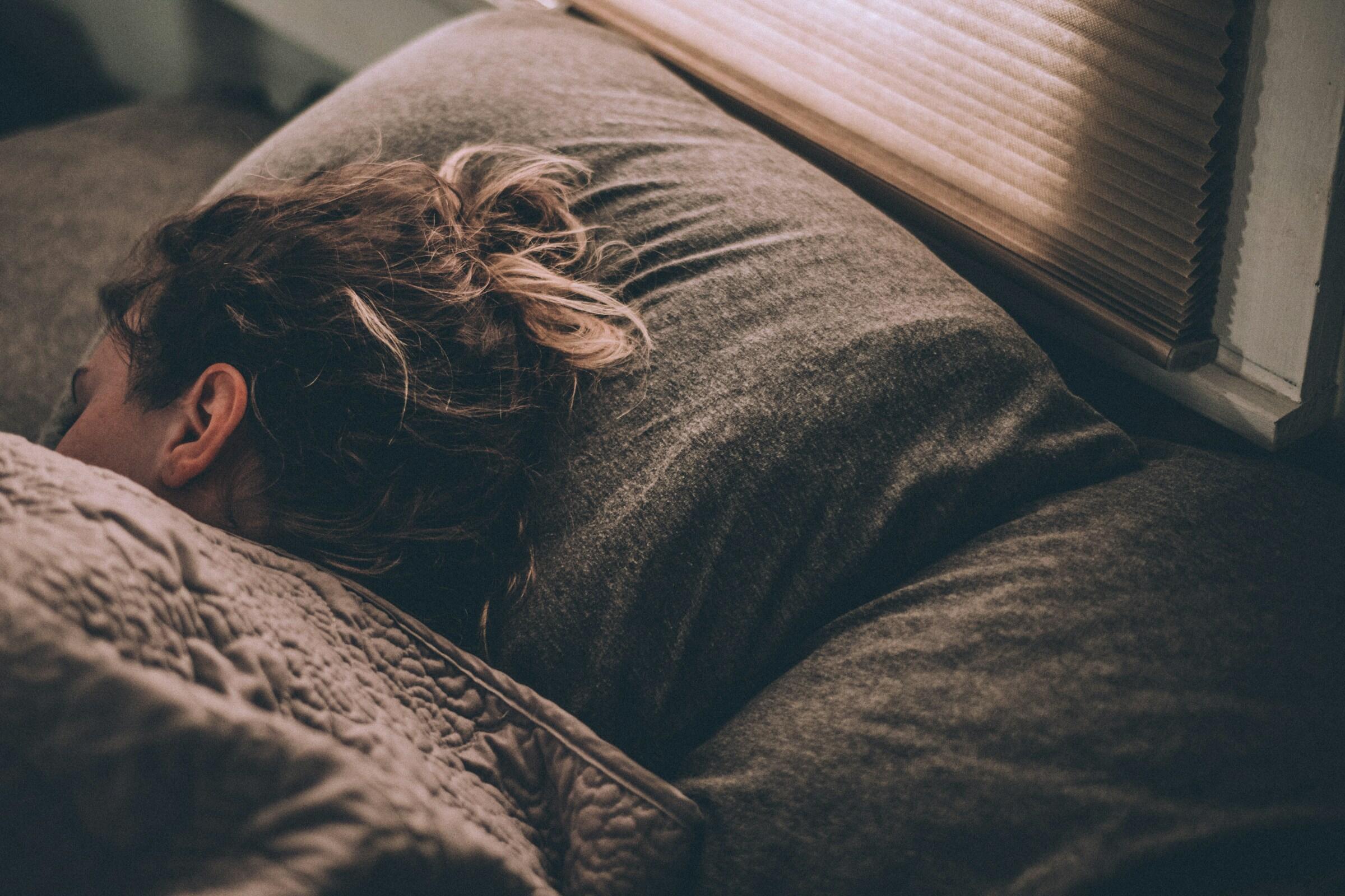Una persona de pelo rizado duerme plácidamente sobre almohadas grises, parcialmente cubierta por una manta con textura, en una habitación suavemente iluminada.