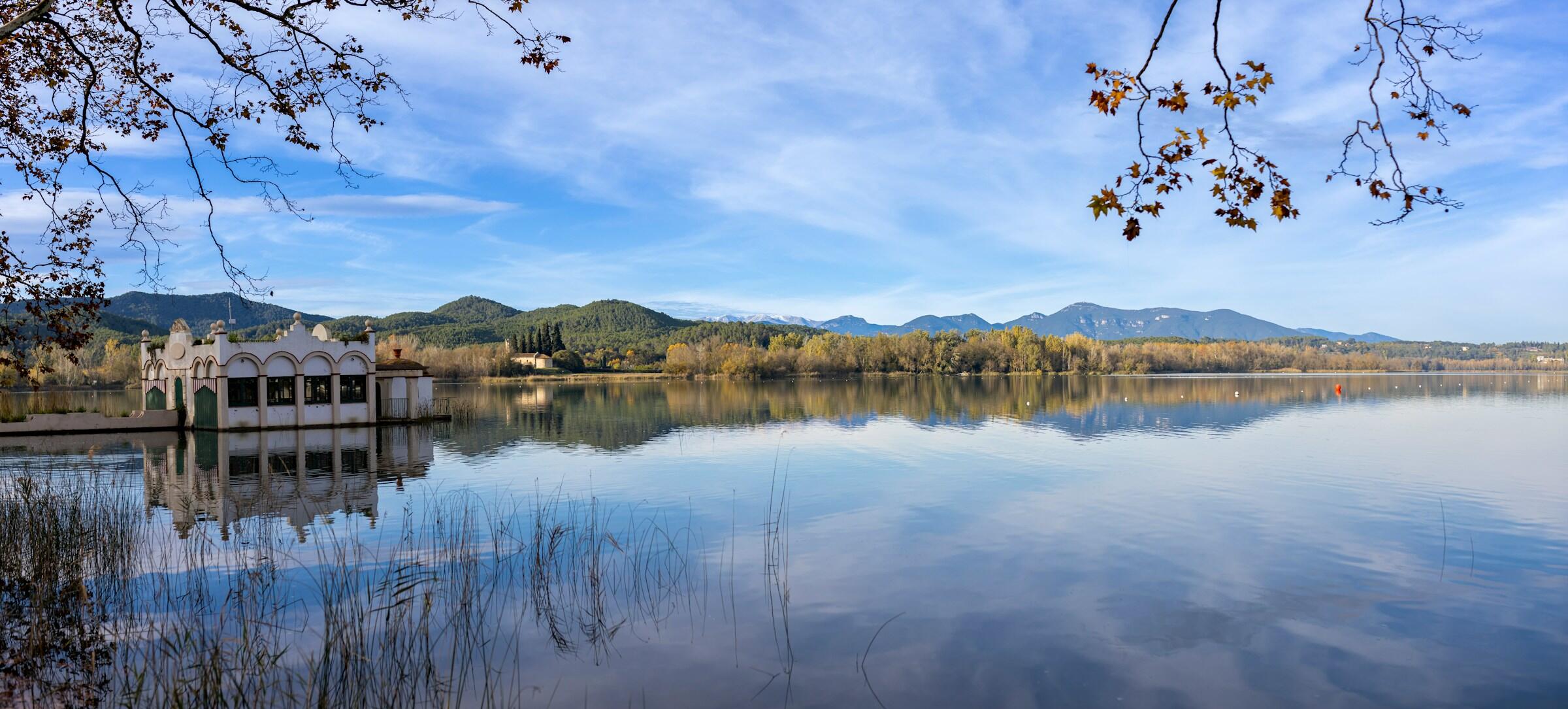 Una tranquila escena lacustre con reflejos de montañas lejanas, un histórico cobertizo para botes y follaje otoñal bajo un cielo azul despejado en el Estany de Banyoles
