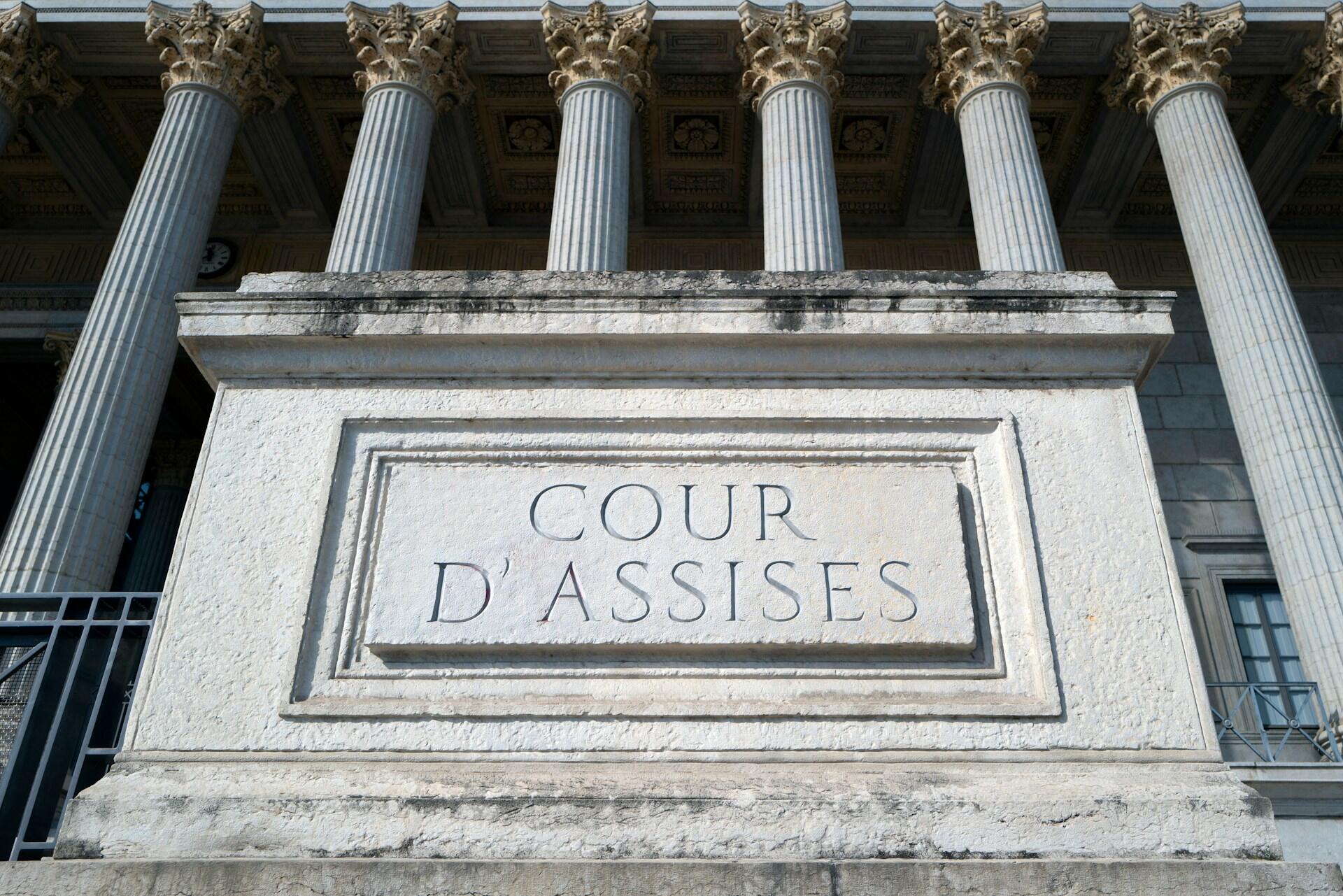Primer plano de un letrero de piedra con la inscripción «COUR D'ASSISES», enmarcado por columnas clásicas de un edificio grandioso. La luz del sol ilumina la textura.