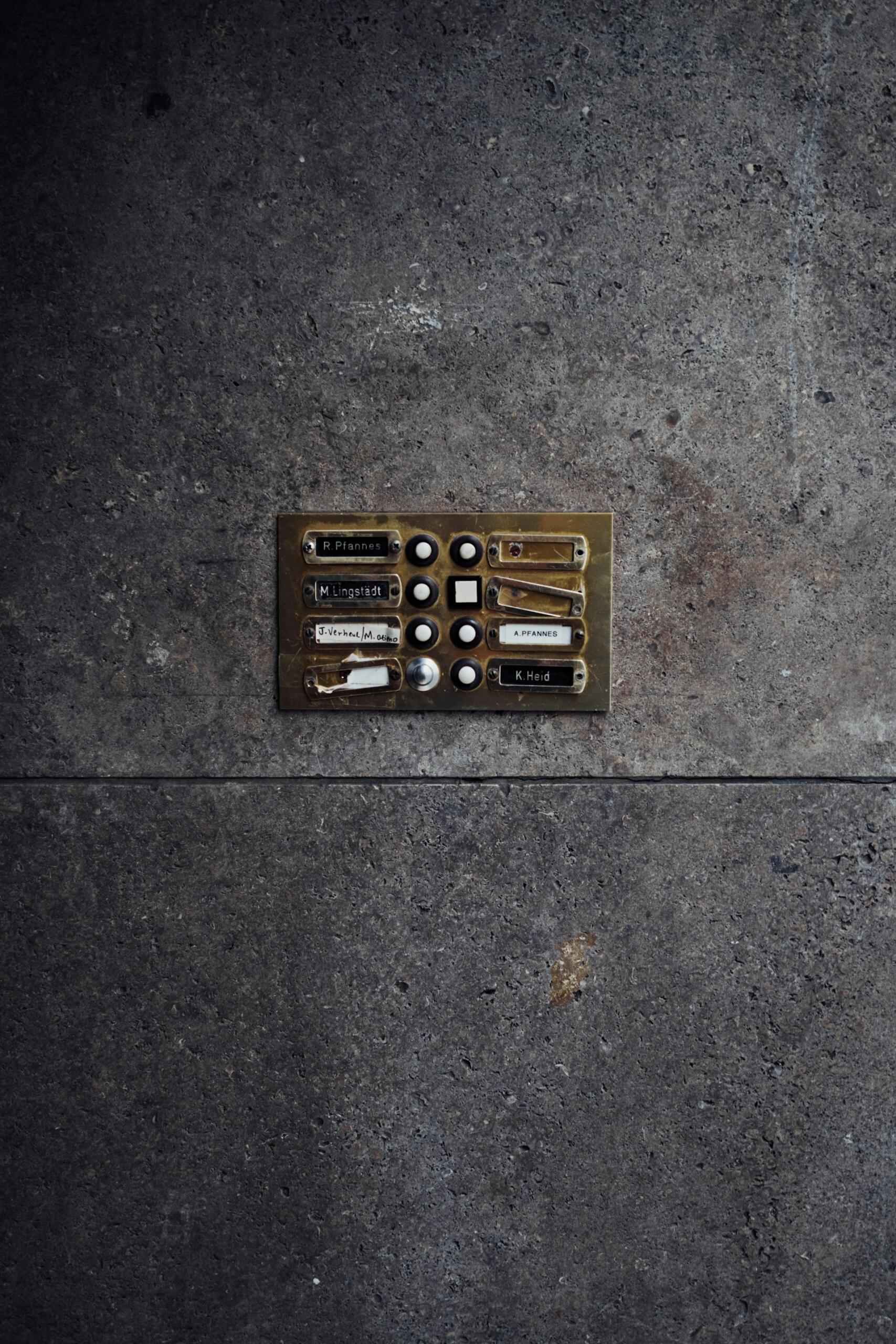 Un interfono desgastado en una pared de piedra gris y rugosa, con seis placas identificativas y botones, que sugiere la entrada de un antiguo bloque de pisos o edificio. Tono urbano y descarnado.