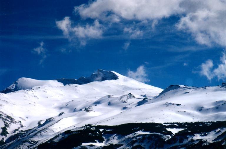 el pico veleta en sierra nevada granada españa cubierto de nieve bajo un cielo azul brillante, con nubes dispersas y laderas rocosas visibles al fondo.