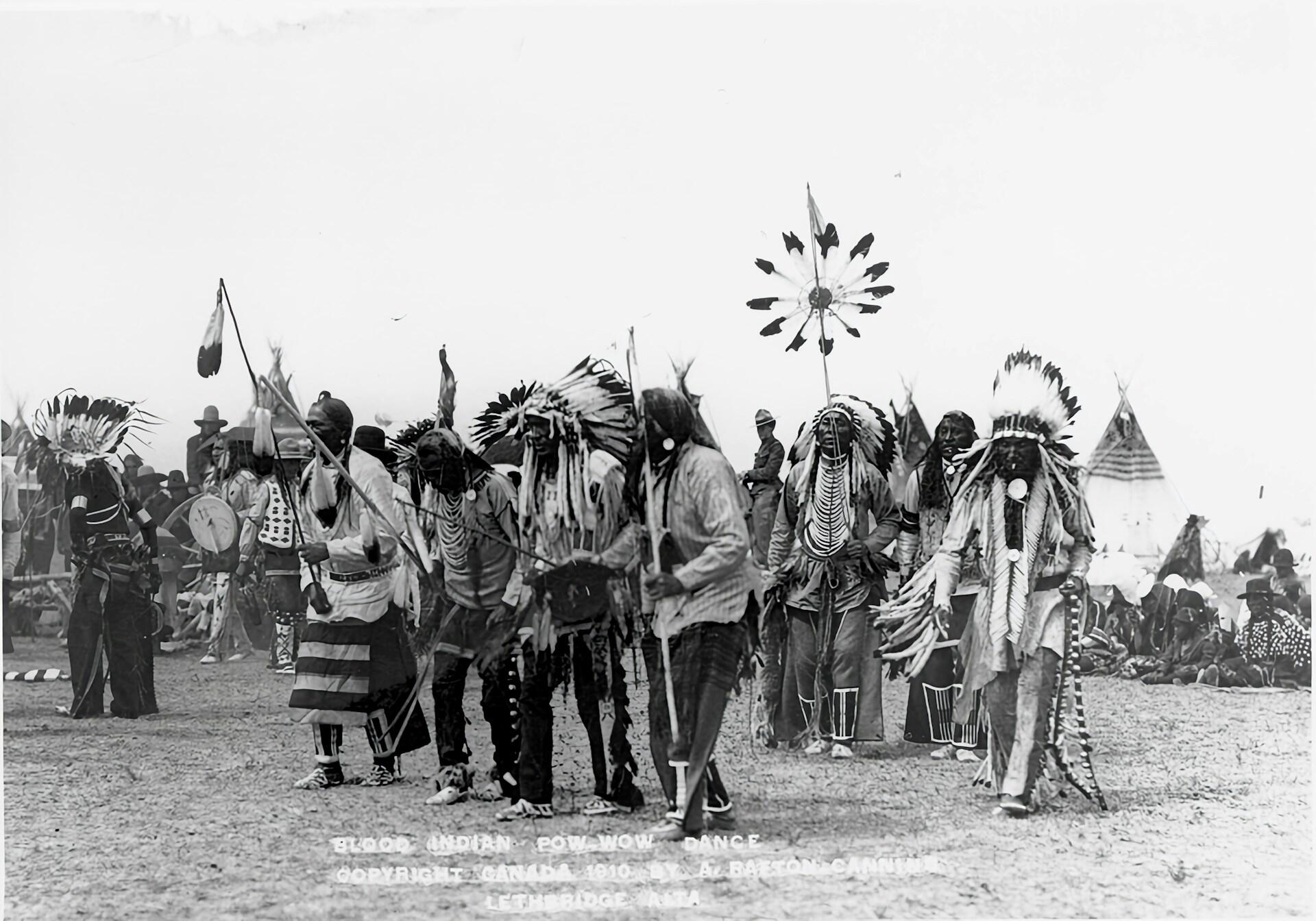 Un grupo de bailarines indígenas con trajes tradicionales participando en un powwow, rodeados de espectadores y tipis.