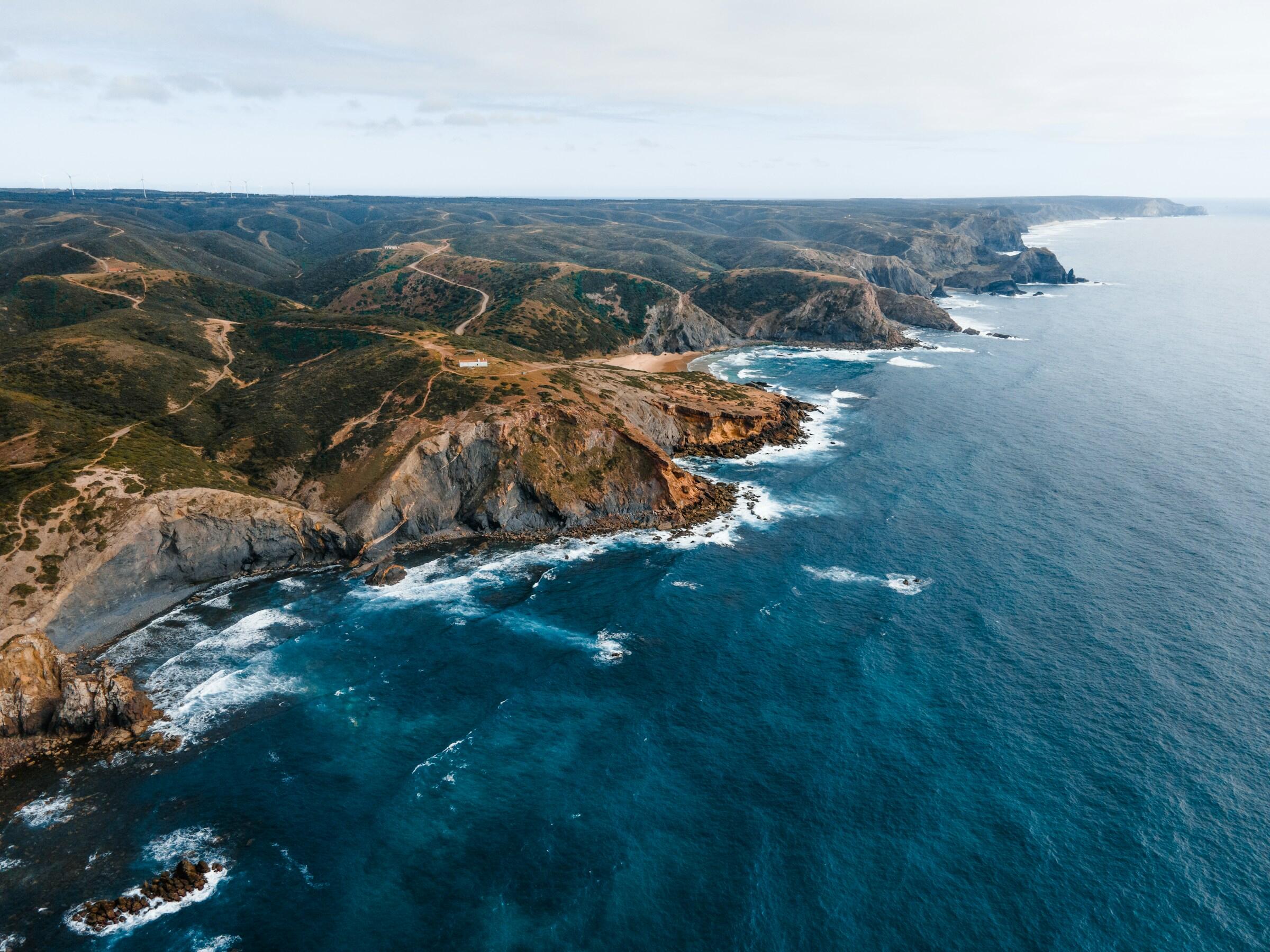 Vista aérea de escarpados acantilados costeros que se encuentran con el océano atlantico azul, rodeados de verdes colinas y un cielo nublado al fondo.