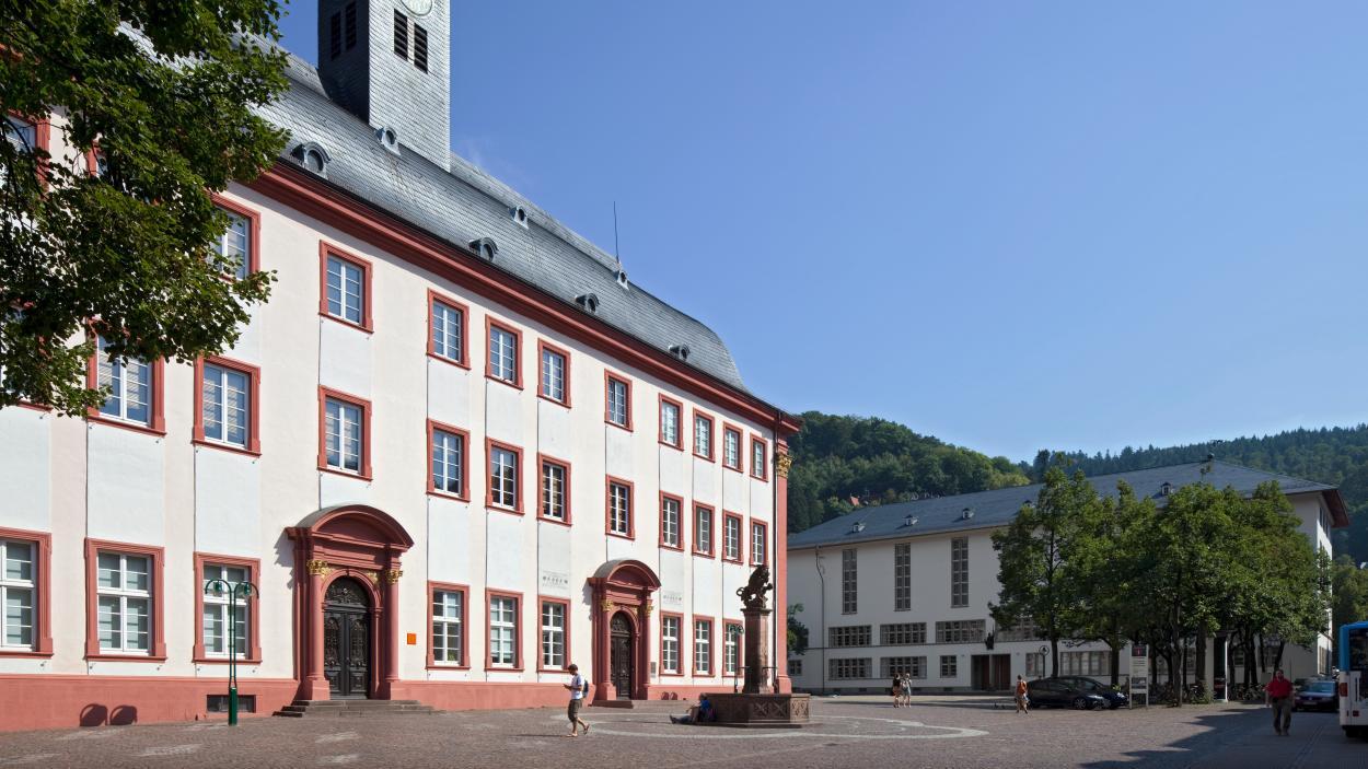 Una plaza histórica europea cuenta con un majestuoso edificio blanco con detalles en rojo, ventanas en arco y una torre del reloj. Árboles y colinas rodean la escena bajo un cielo azul y despejado.
