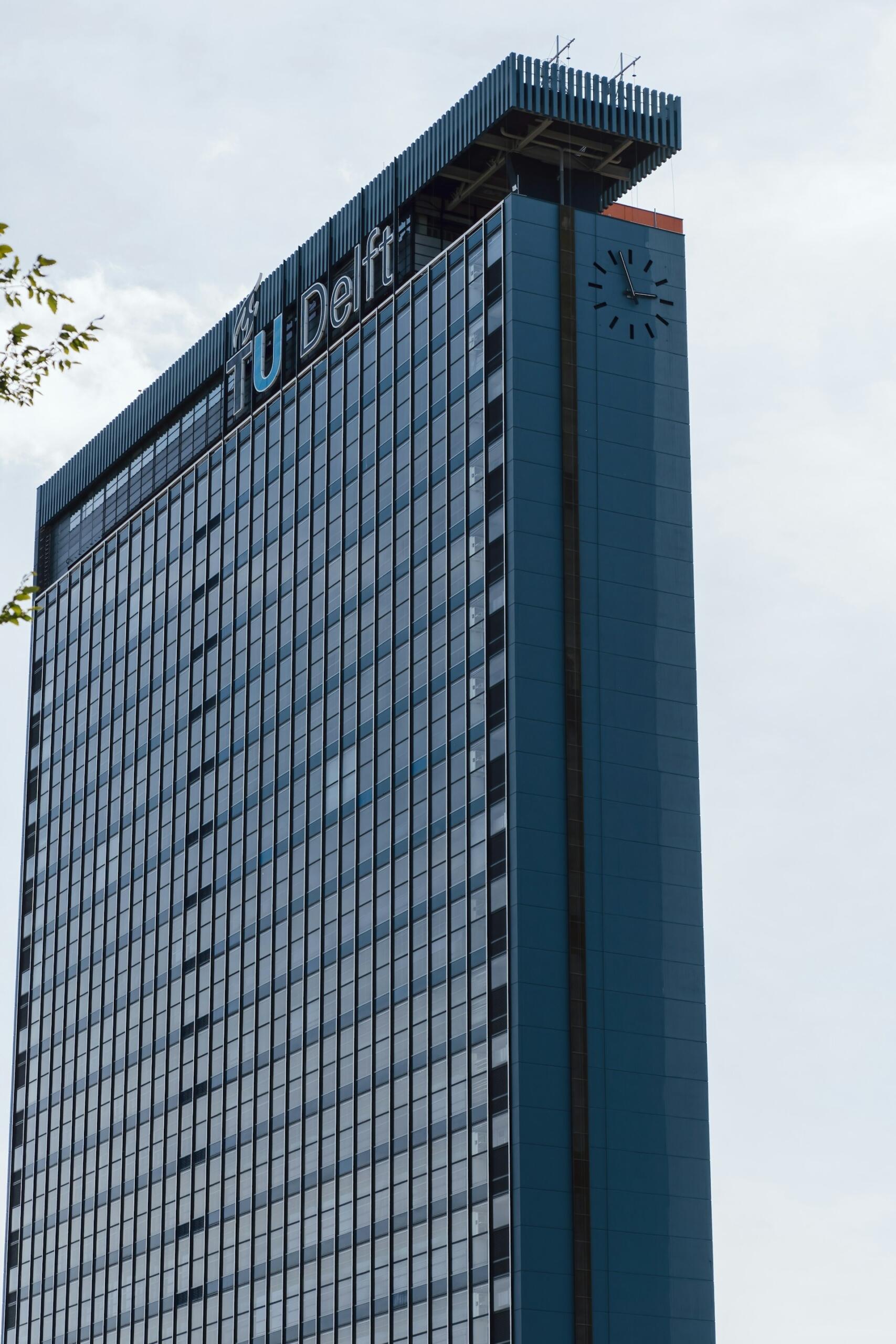 Un edificio alto y moderno con ventanas de cristal reflectante y el letrero «TU Delft» cerca de la parte superior. Se ve un reloj, con un cielo nublado de fondo.