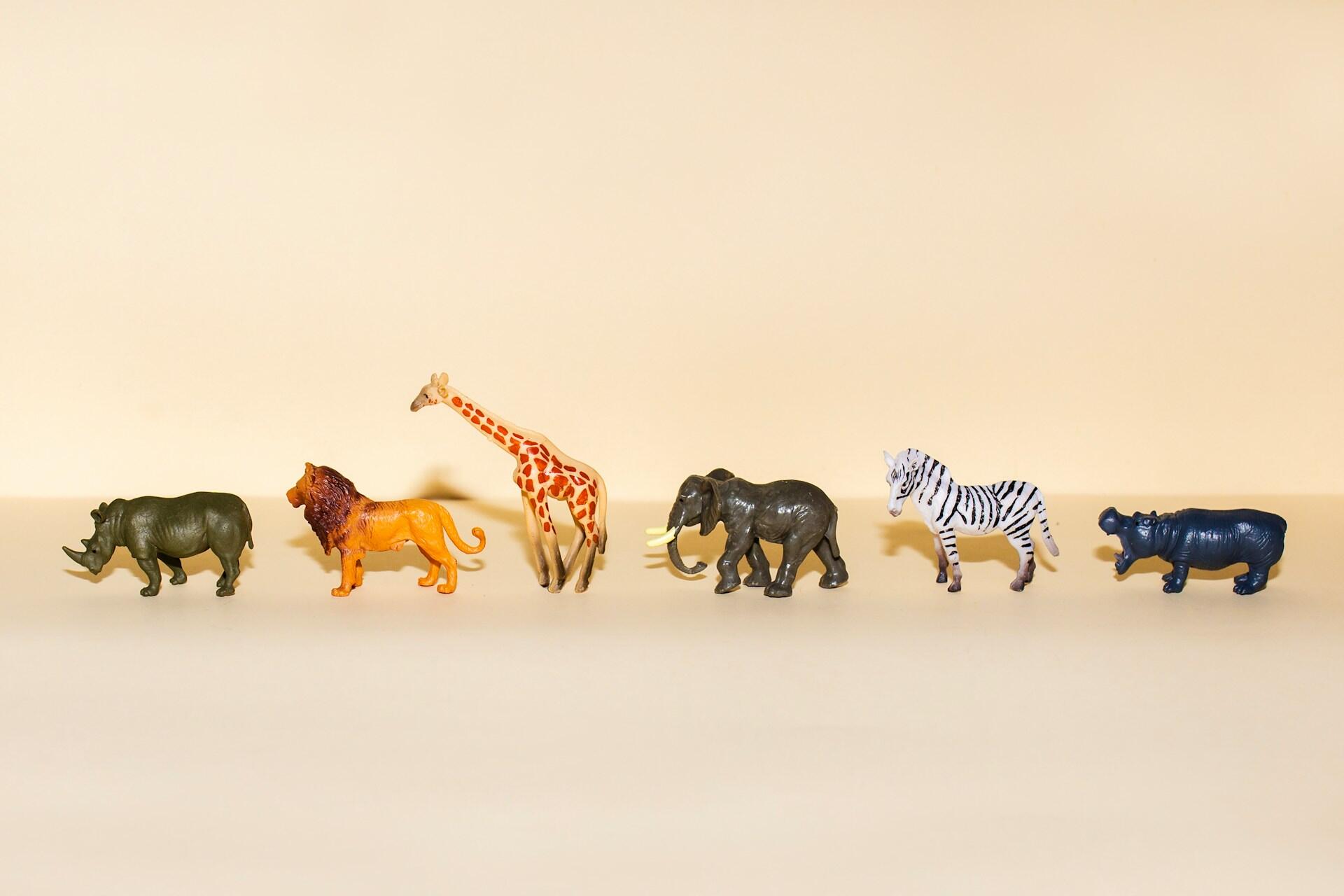 Una fila de coloridos animales de juguete, entre los que se incluyen un rinoceronte, un león, una jirafa, un elefante, una cebra y un hipopótamo, sobre un fondo beige suave.