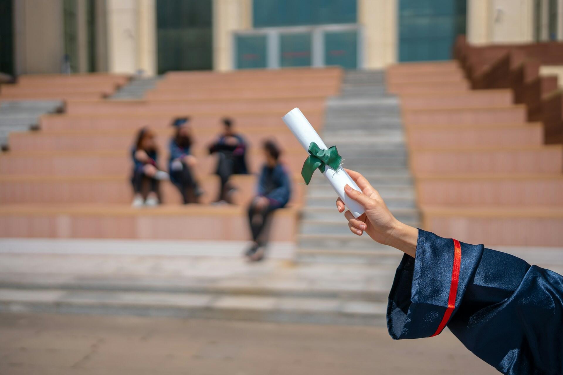 Un graduado con toga azul sostiene con orgullo un diploma enrollado con una cinta verde, mientras sus amigos celebran en el fondo.