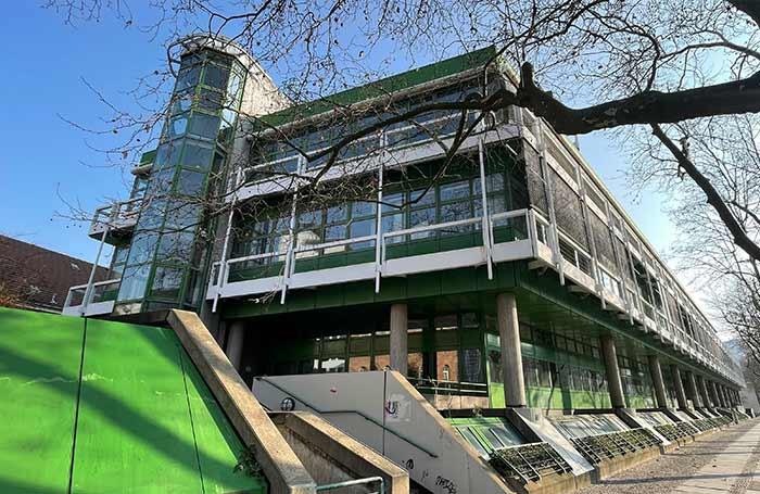 Un edificio moderno y de líneas angulosas, con grandes ventanales y toques de verde, se alza bajo las ramas desnudas de los árboles, con un cielo azul claro de fondo, transmitiendo una sensación de elegancia urbana.