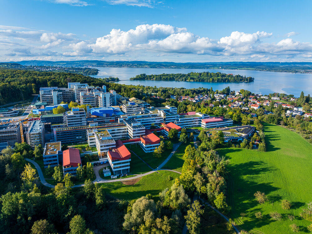 Vista aérea de un campus universitario amplio y moderno, con edificios de tejados rojos, rodeado de una exuberante vegetación, junto a un lago tranquilo bajo un cielo azul.