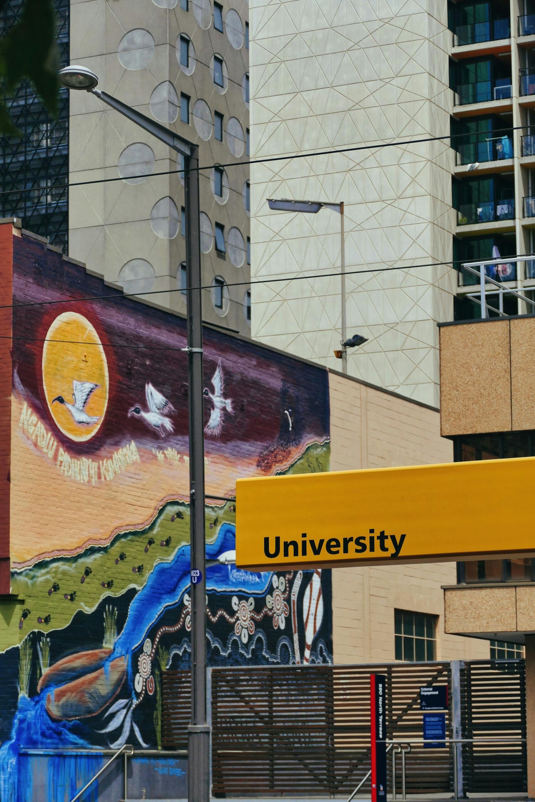 Un mural lleno de vida, con pájaros volando y un sol poniente, adorna una pared urbana. Un letrero de color amarillo intenso en el que se lee «Universidad» contrasta con las fachadas de los edificios modernos.